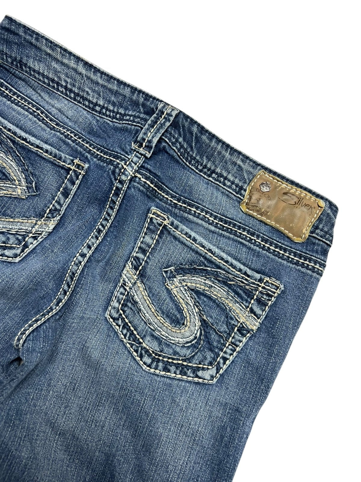 Eine Nahaufnahme der Silver Jeans Low Rise Boot von ZAZA resell in Größe 29 (M). Sie zeigt blauen Denim mit aufgestickten Taschendesigns, einen braunen Leder-Markenaufnäher am Bund und Bundweite-Details für eine perfekte Passform.