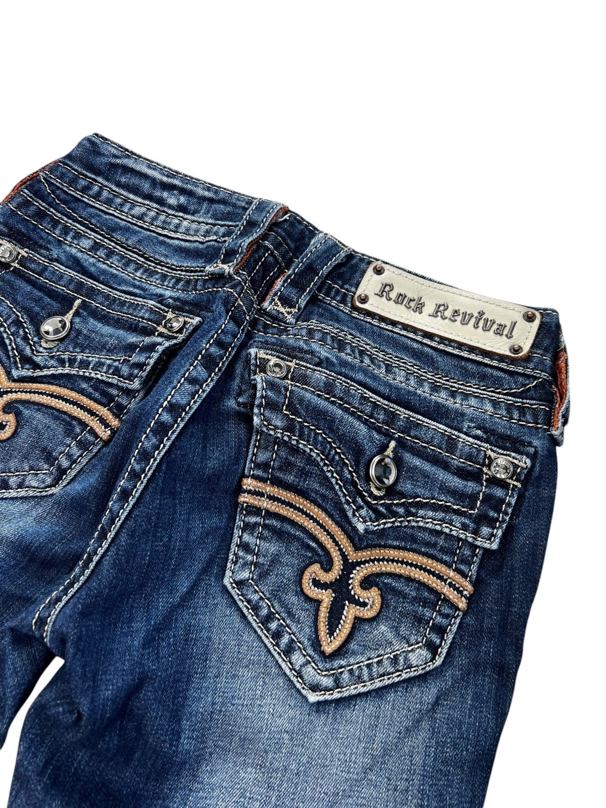 Eine Nahaufnahme der Rückseite der von ZAZA weiterverkauften Rock Revival Low Rise Boot Jeans (W24 x L30) in Blau, die dekorative Nähte, silberne Knöpfe, ein Markenetikett über der rechten Tasche und eine detaillierte Beinöffnung zeigt.