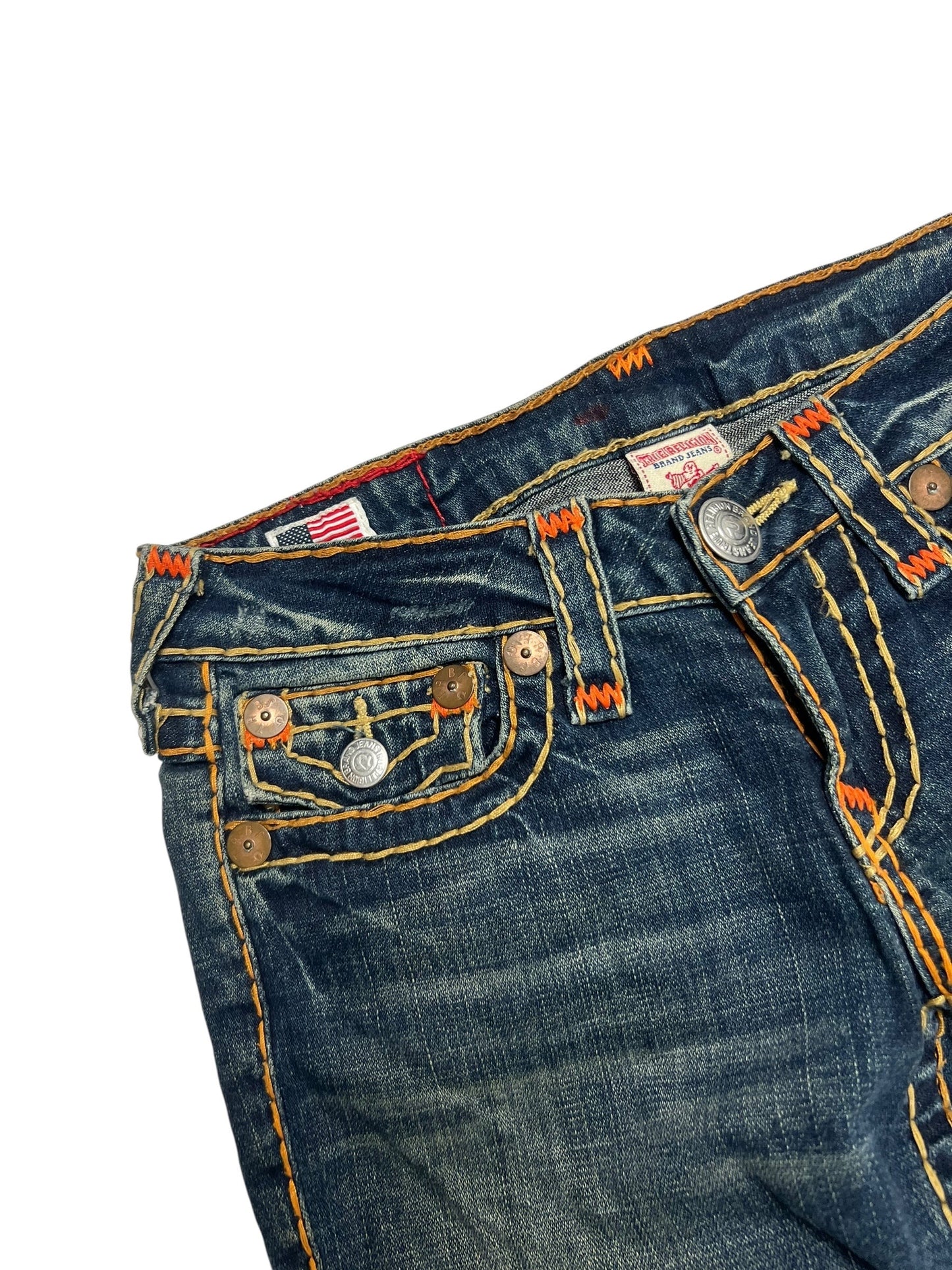 Eine Nahaufnahme von Bund und Tasche der True Religion Billy Super T - Low Rise Straight | 27 (M) Jeans von ZAZA resell, mit dunkelblauem Denim, orangefarbenen Nähten, Used-Details, Bundweite-Etikett, Metallknöpfen und einer amerikanischen Flagge auf weißem Hintergrund.