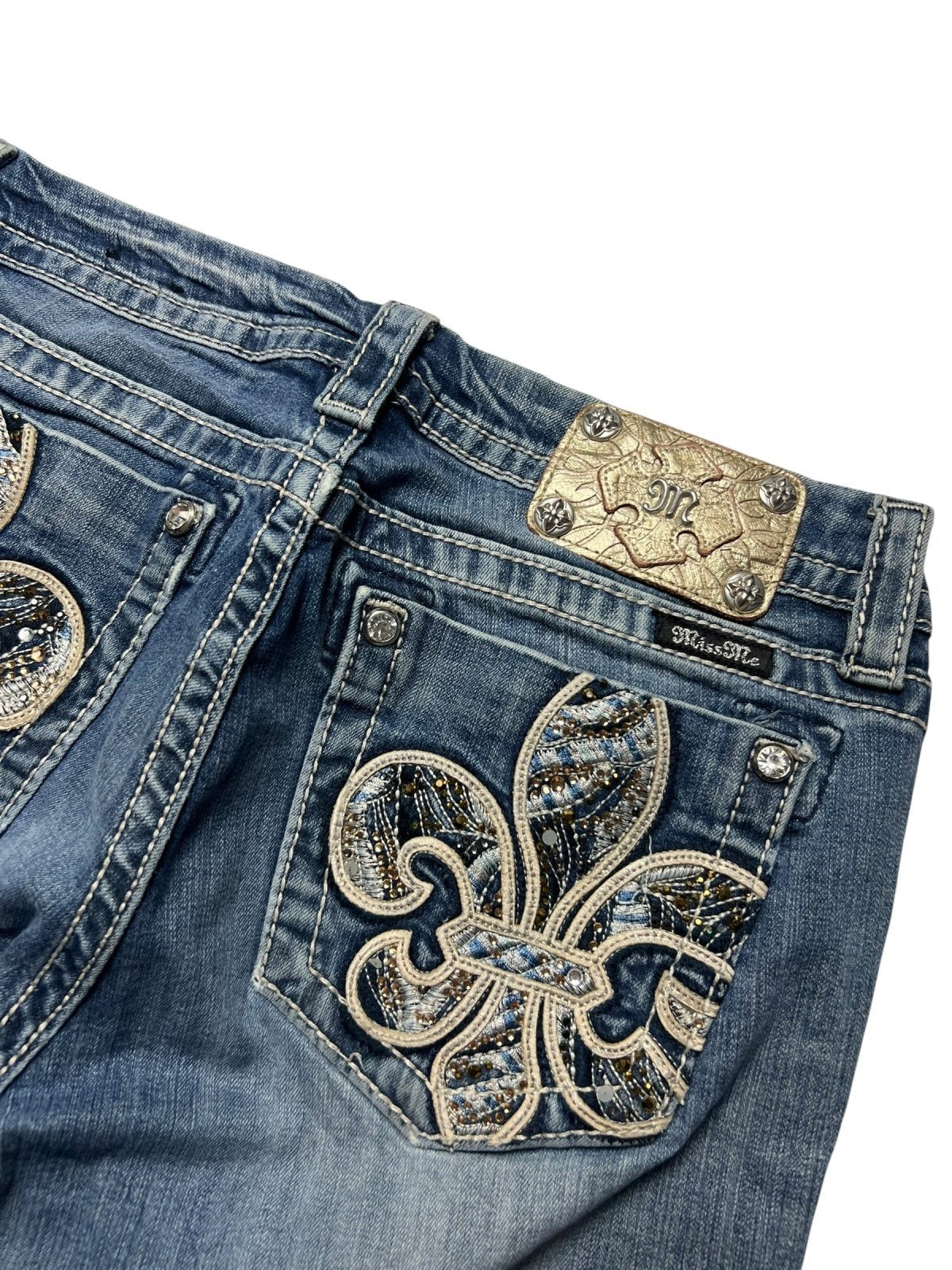 Eine Nahaufnahme der blauen Denim-Jeans Miss Me Mid Rise Boot (Größe 30/M) von ZAZA resell, mit einem verzierten Fleur-de-Lis-Strassstein-Design auf der Gesäßtasche und einem dekorativen Metall-Label über der Tasche für eine perfekte Passform.