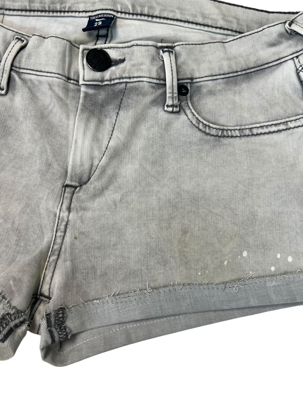 Eine Nahaufnahme der True Religion Shorts | 29 (M) von ZAZA resell aus hellgrauem Denim mit ausgefransten Säumen, schwarzem Knopf, Frontreißverschluss und schwachen Flecken sowie kleinen weißen Flecken am unteren Teil.