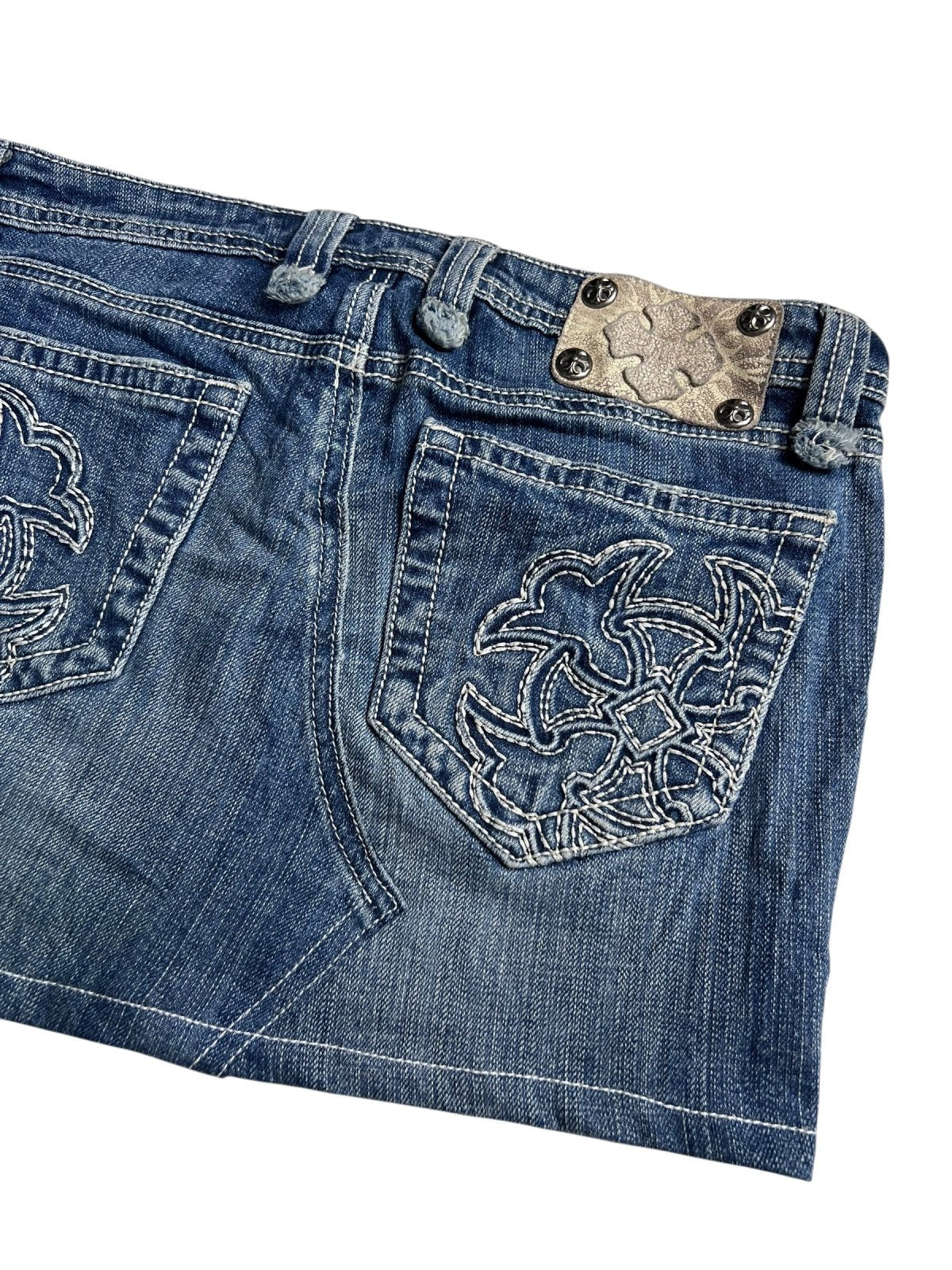 Eine Nahaufnahme des Miss Me Skirt | 28 (M) von ZAZA resell zeigt blauen Denim mit weiß bestickten Gesäßtaschen und einem Metallic-Patch in der Nähe des Bundes, der Details wie Bundweite und Außenbeinlänge für eine individuelle Rock-Passform hervorhebt.