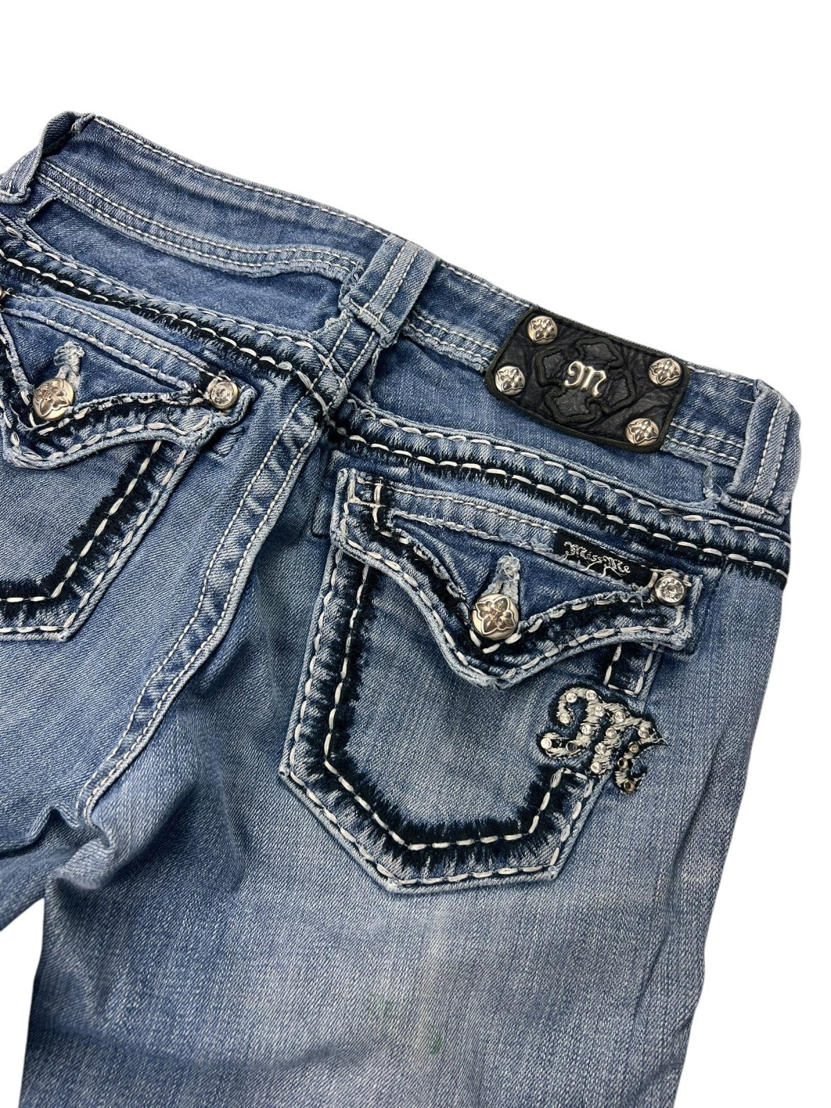 Eine Nahaufnahme der Miss Me Mid Rise Boot Jeans (W26 x L29) von ZAZA Resell. Sie zeigt blauen Denim mit dicken weißen Nähten, dekorative Gesäßtaschen mit silbernen Knöpfen, ein gesticktes M und einen schwarzen Lederaufnäher mit metallischen Akzenten in der Nähe des Hosenbunds.