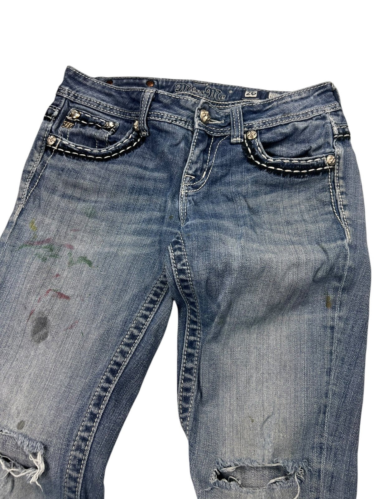 Die Miss Me Mid Rise Boot Jeans von ZAZA Resell (W26 x L29) besteht aus blauem Distressed-Denim mit zerrissenen Knien, Farbflecken, Ziernähten, Metallnieten an den Vordertaschen und einer bequemen Bundweite für eine ideale Passform.