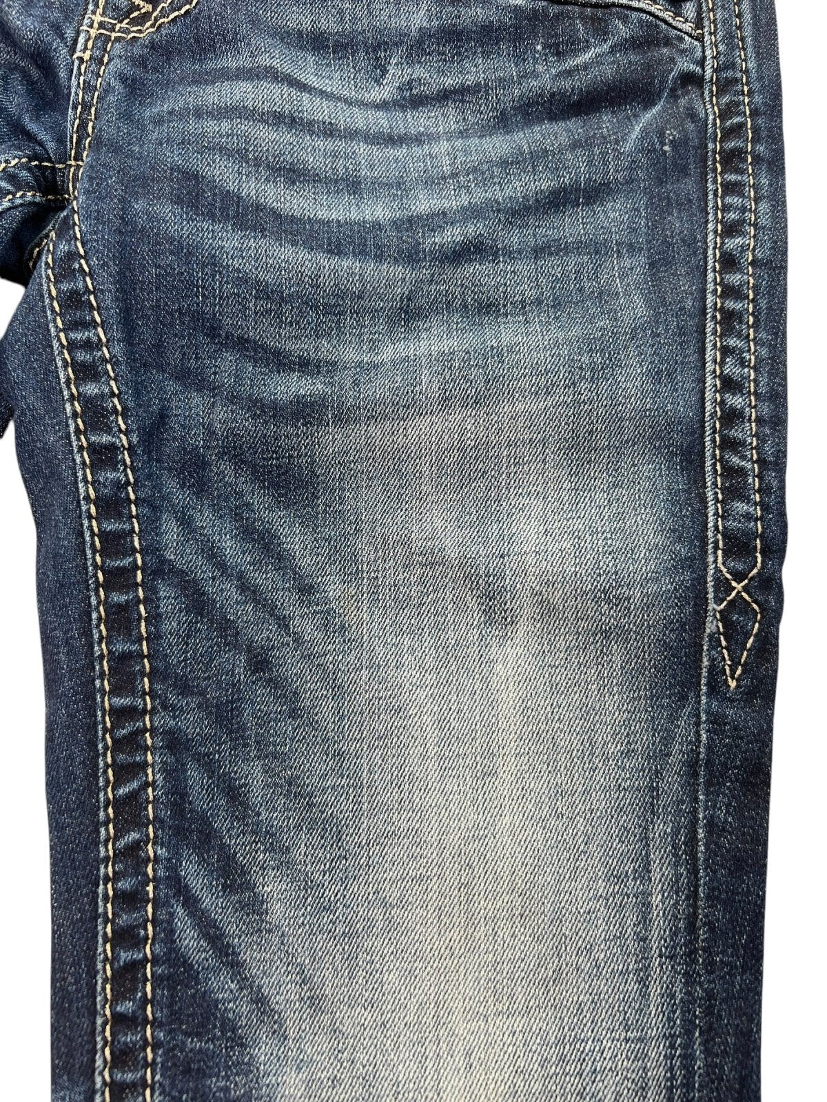 Nahaufnahme der Rock Revival Low Rise Boot Jeans (W24 x L30) von ZAZA Resell, bestehend aus blauem Denim mit verblichenem, abgenutztem Stoff und weißen Nähten, die die Details der Bundweite und Beinöffnung betonen.