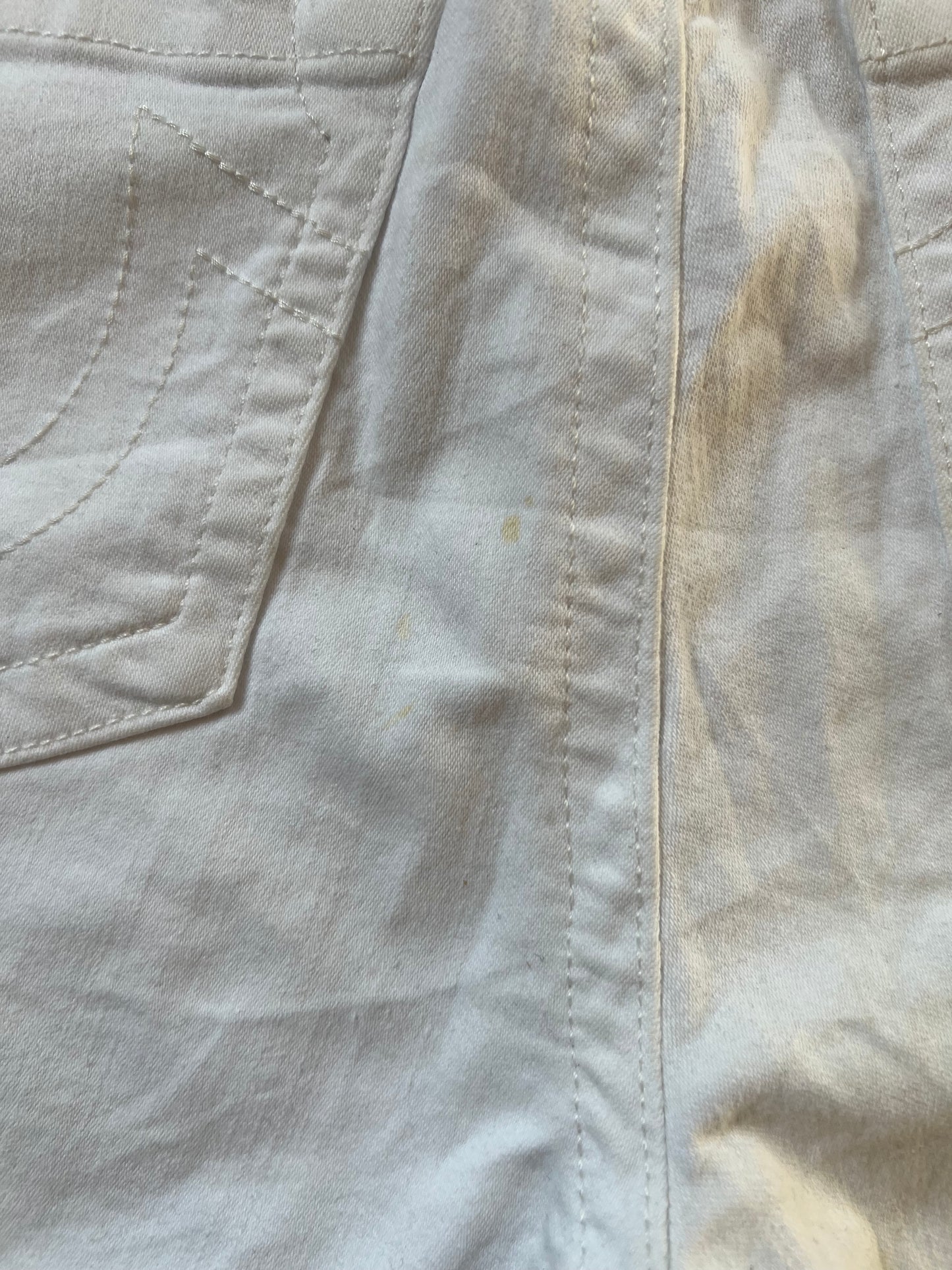 Nahaufnahme der weißen True Religion Mid Rise Shorts (Größe 30, Bundweite 39) von ZAZA resell, die eine linke Tasche und schwache gelbe Flecken in der Nähe der Mittelnaht zeigt. Der Stoff erscheint leicht zerknittert.