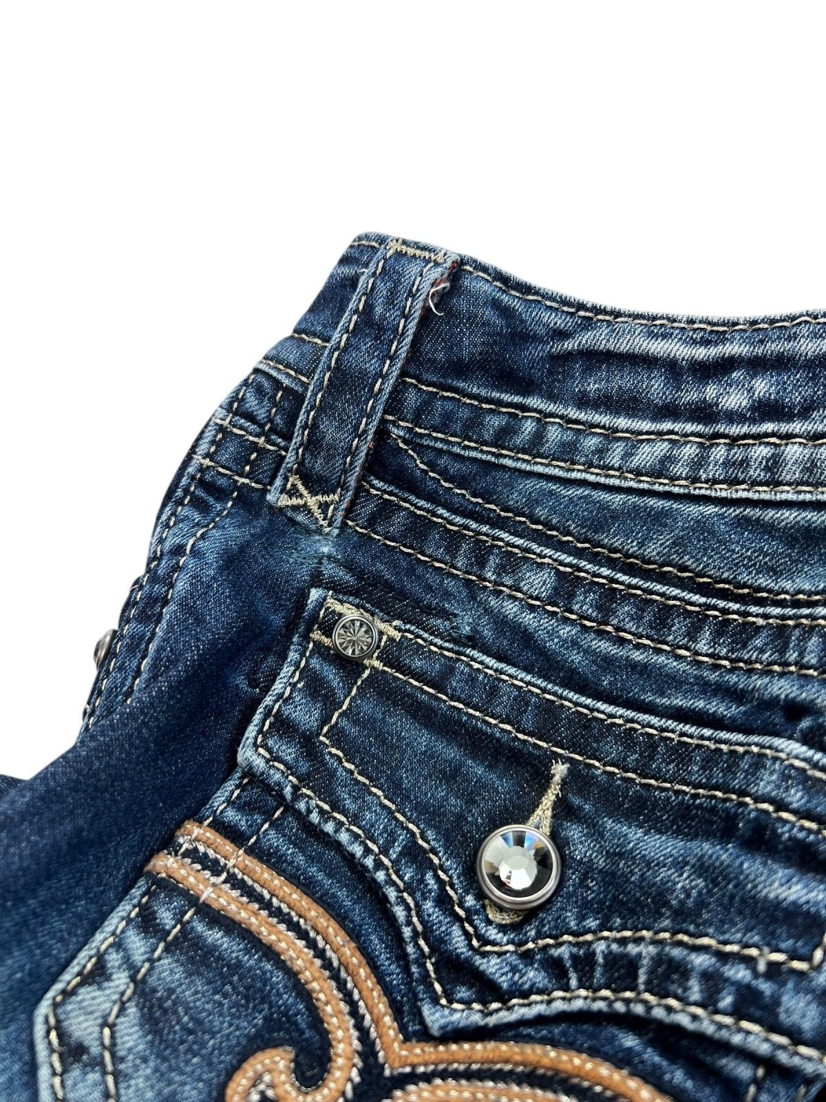 Nahaufnahme des Hosenbunds und des Taschenbereichs der von ZAZA weiterverkauften Rock Revival Low Rise Boot Jeans (B24 x L30) aus blauem Denim mit Ziernähten, silbernen Nietenknöpfen und detaillierter weißer und hellbrauner Stickerei. Weißer Hintergrund.
