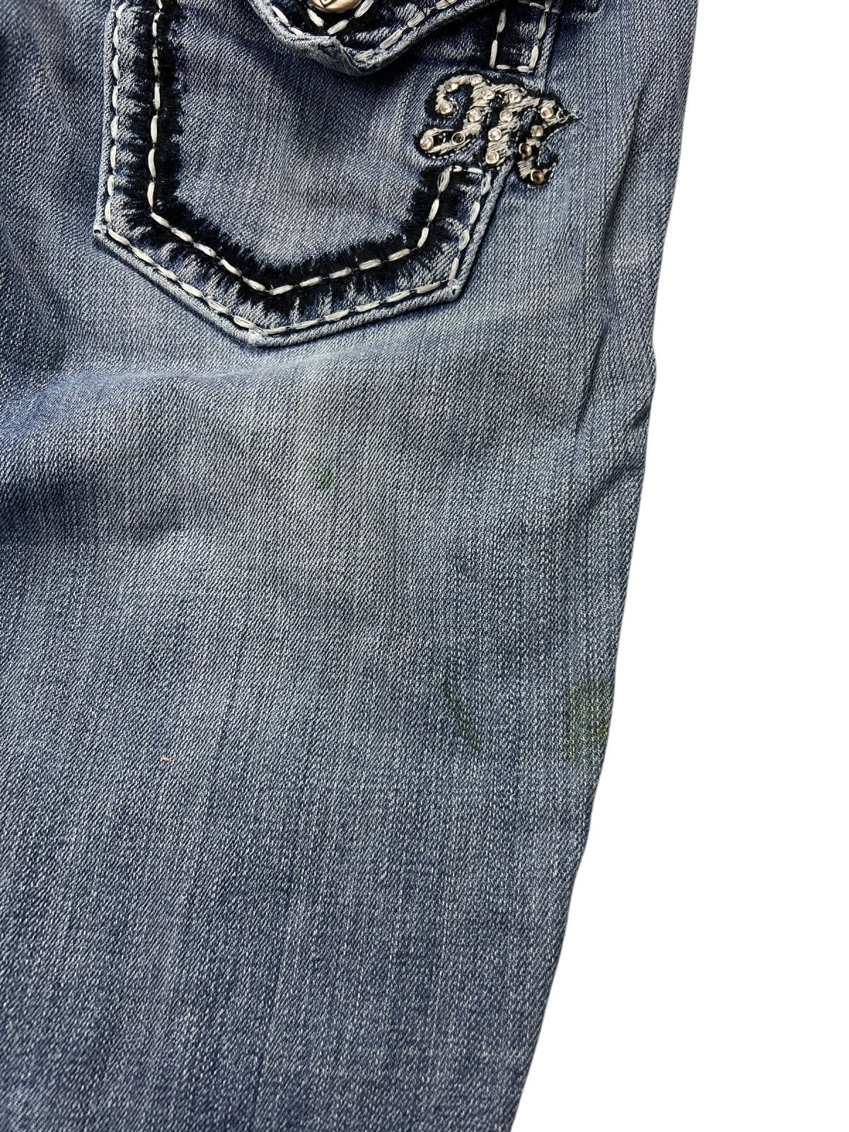 Nahaufnahme der Miss Me Mid Rise Boot Jeans von ZAZA Resell (W26 x L29) in blauem Denim, mit aufgenähter Gesäßtasche mit dekorativem „M“-Emblem, schwachen grünen Flecken unter der Tasche und präziser Beinöffnung für verbesserte Passform und Stil.