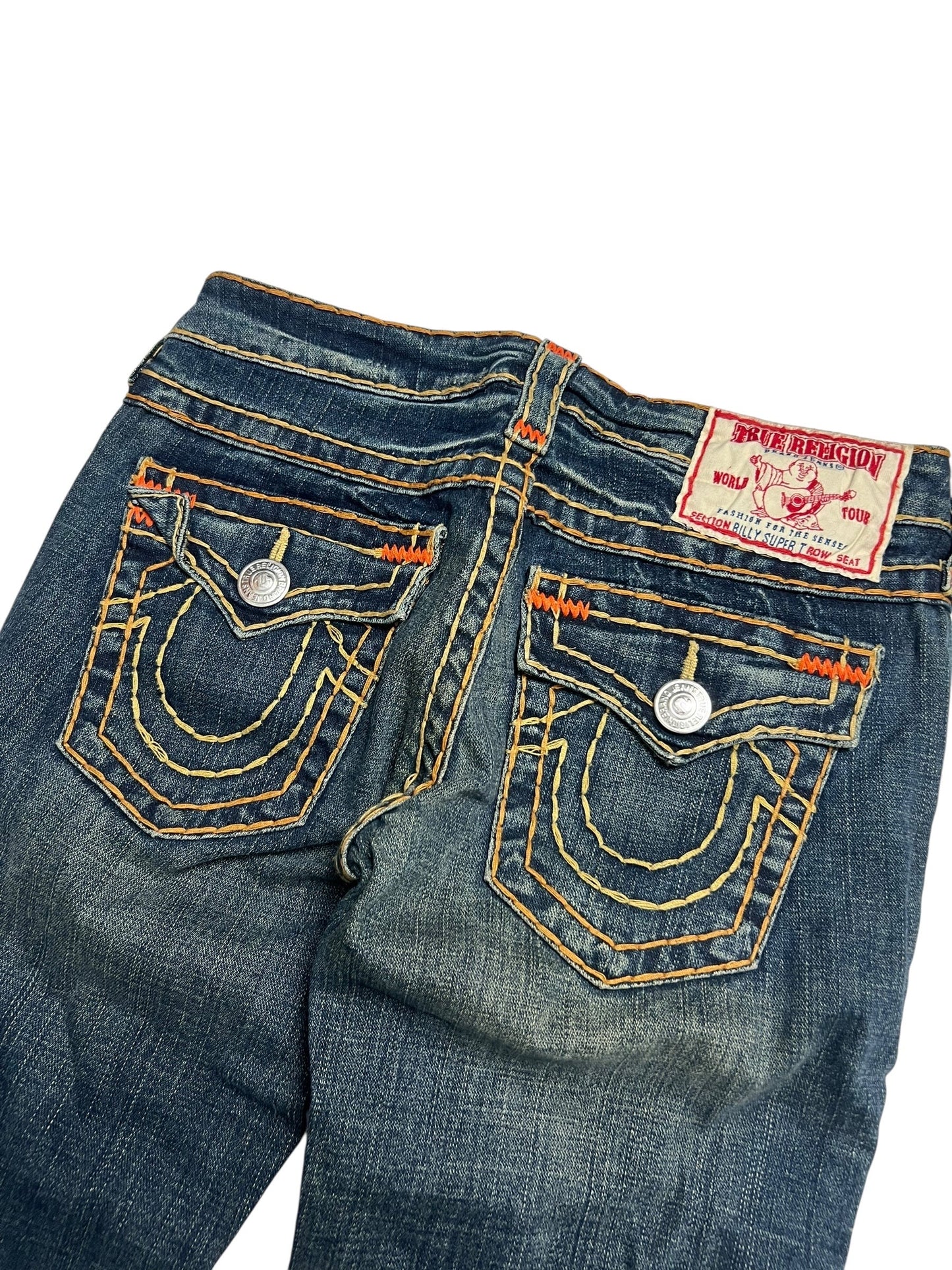 Eine Nahaufnahme der True Religion Billy Super T Jeans von ZAZA resell in Größe 27 (M) zeigt blauen Denim, gelb-orangefarbene Nähte, zwei detaillierte Gesäßtaschen, silberne Knöpfe und ein rot-weißes Label, das Bundweite und gekürzte Außenbeinlänge hervorhebt.