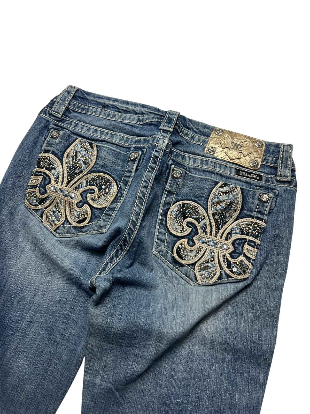 Die Miss Me Mid Rise Boot Jeans von ZAZA resell in Größe 30 (M) hat verzierte Gesäßtaschen mit großen silbernen und goldenen Fleur-de-Lis-Motiven, Strasssteinen, Ziernähten und einem metallischen Markenaufnäher über der rechten Tasche.