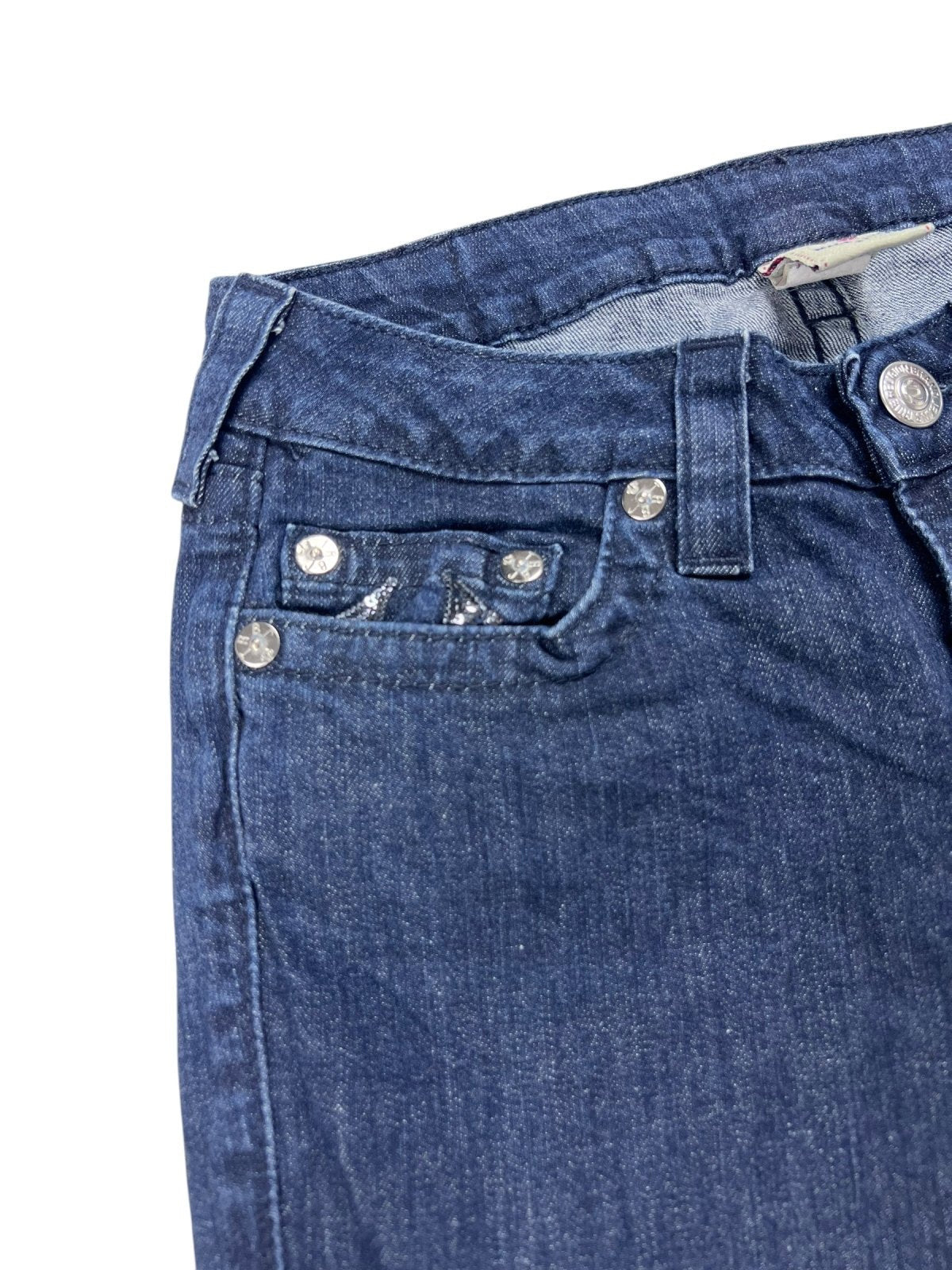 Die Nahaufnahme des oberen Teils der ZAZA-Reseller-Jeans True Religion Low Rise Boot (B27 x L34) zeigt Bund, Gürtelschlaufen, Knopf, Vorderseite und Münztasche mit Nieten. Der blaue Denim hat eine leicht abgenutzte Textur und ist mit einem Etikettgröße-Label versehen.