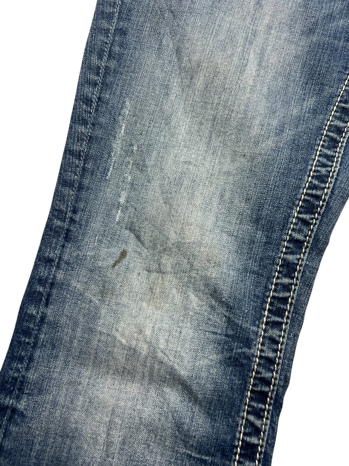 Eine Nahaufnahme der Miss Me Mid Rise Boot Jeans von ZAZA resell in Größe 30 (M) zeigt verblasstes blaues Denim mit Schmutzflecken, weiße Nähte entlang der Naht und den Bundweite-Schriftzug in der Nähe des Bunds.