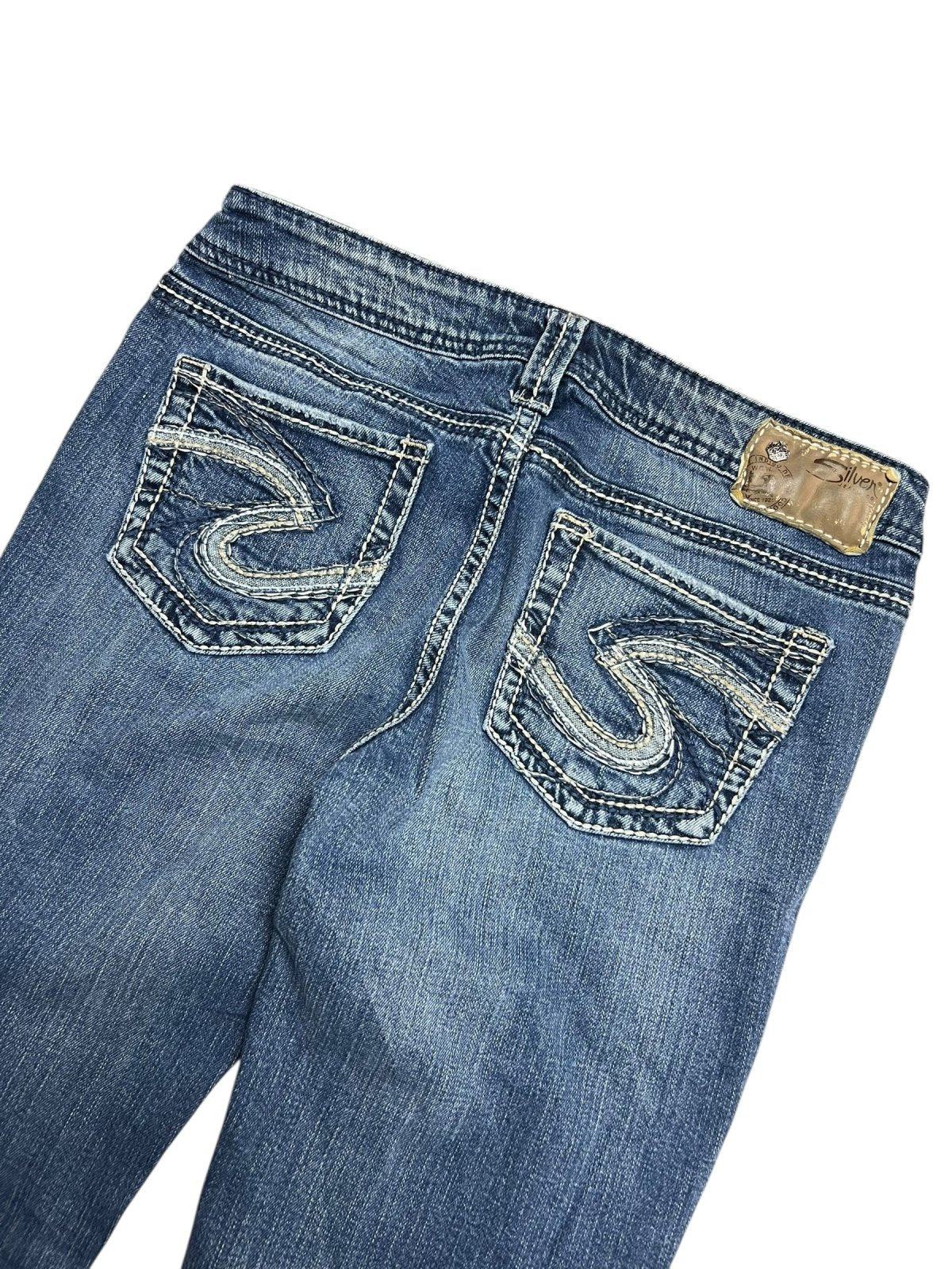 Die Silver Jeans Low Rise Boot von ZAZA resell in Größe 29 (M) ist eine blaue Jeans in Rückenansicht mit zwei weiß bestickten Gesäßtaschen, einem hellbraunen Lederbundaufnäher und einem Etikett mit Bundweite und Labelgröße.