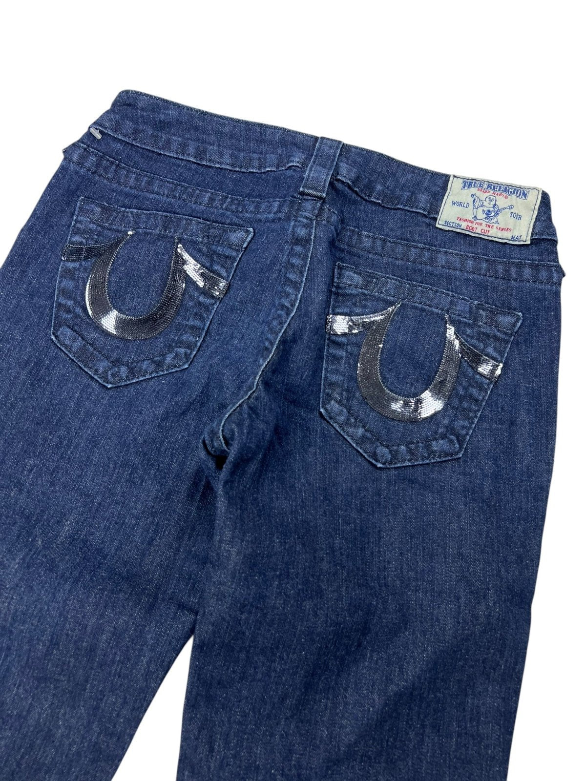 Eine blaue True Religion Low Rise Boot Jeans (W27 x L34) von ZAZA Resell, von hinten gezeigt, mit charakteristischen Pailletten-Hufeisen-Nähten auf beiden Taschen und einem Marken-Bundetikett. Etikettgröße als Referenz verfügbar.