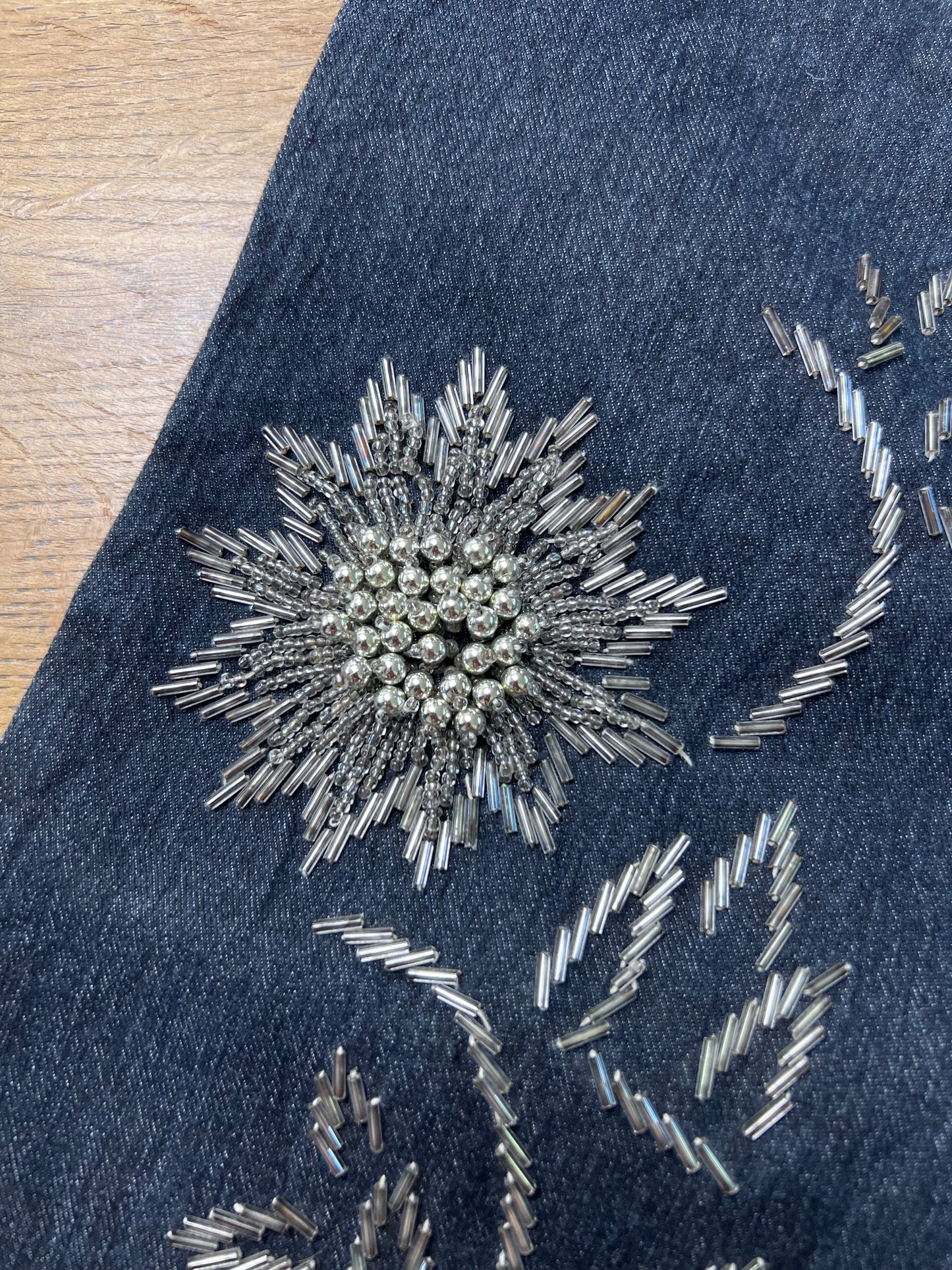 Nahaufnahme des Denim-Schuhs „Edith Mid Rise Boot“ (B26 x L32) von ZAZA. Er zeichnet sich durch eine auffällige, silberne Blumenverzierung mit runden Perlen und röhrenförmigen Blütenblättern in der Nähe der Beinöffnung aus. Im Hintergrund ist eine Holzoberfläche zu sehen.