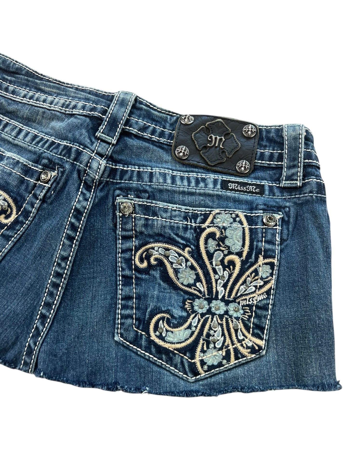 Eine Nahaufnahme des Miss Me Skirt | W28 von ZAZA Resell zeigt blauen Denim mit Ziernähten, einer gestickten Lilie auf der Gesäßtasche und einem schwarzen Miss Me-Aufnäher aus Leder – ein herausragendes Einzelstück.