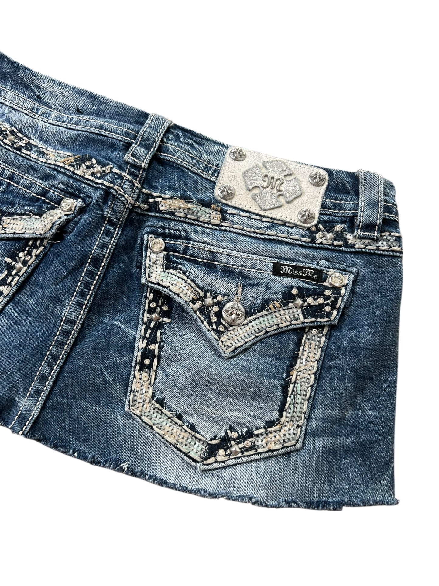 Zugeschnittenes Bild des Miss Me-Rocks von ZAZA Resell, einem Minirock aus blauem Denim mit Ziernähten, Strasssteinen, einem silberverzierten Patch über der Gesäßtasche und detaillierter Stickerei mit metallischen Akzenten für einen rockigen Look.