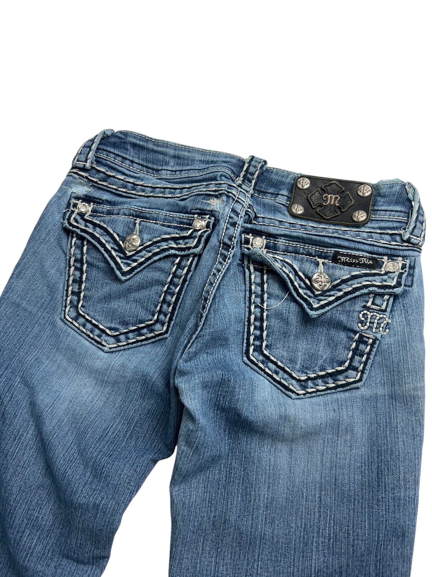 Der Miss Me Low Rise Boot (W25 x L30) von ZAZA Resell präsentiert sich in blauem Denim, mit weißen Nähten, aufgenähten, verzierten Gesäßtaschen mit silbernen Knöpfen, einem schwarzen Lederpatch am Bund und einem dezenten Used-Look.