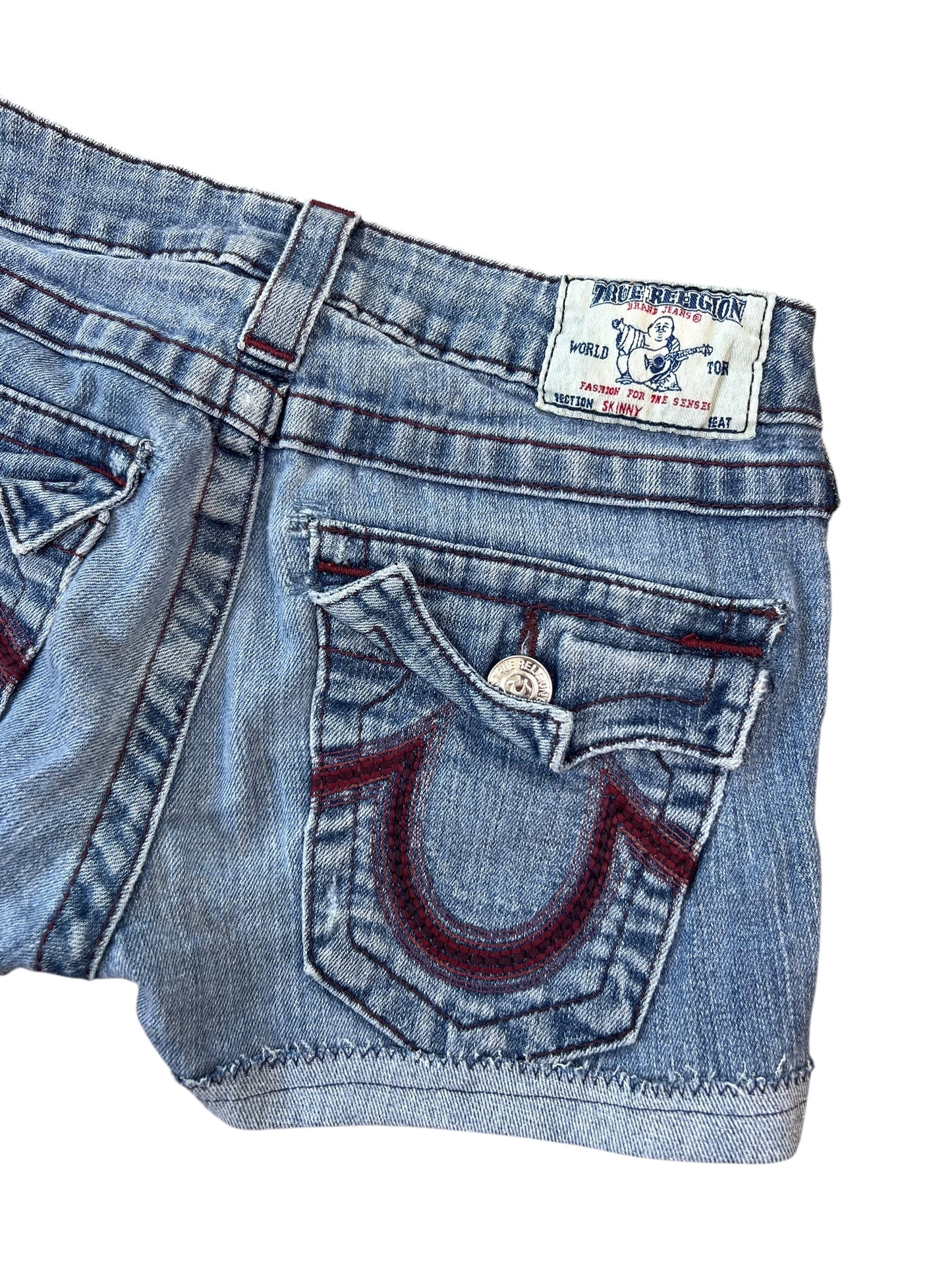Nahaufnahme der blauen True Religion Shorts aus Denim (Größe 25/XS) von ZAZA resell mit Gesäßtasche mit Klappe, roten Nähten, Metallknopf und einem weißen Etikett mit einem Gitarristen über der Tasche.