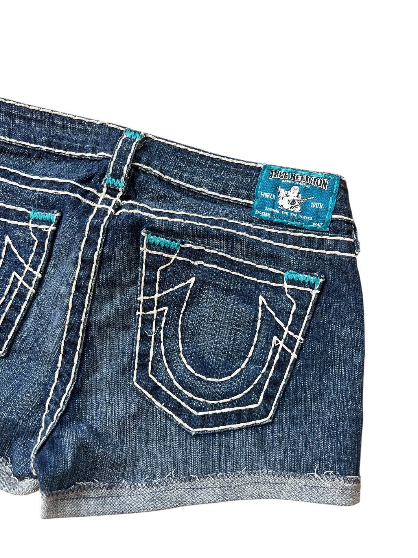 Eine Nahaufnahme der blauen True Religion Shorts (Größe 30/M) von ZAZA resell mit weißen Nähten, ausgefranstem Saum, hufeisenförmigem Detail an der Gesäßtasche und einem blauen Markenetikett über der rechten Tasche. Außenbeinlänge 26 cm.