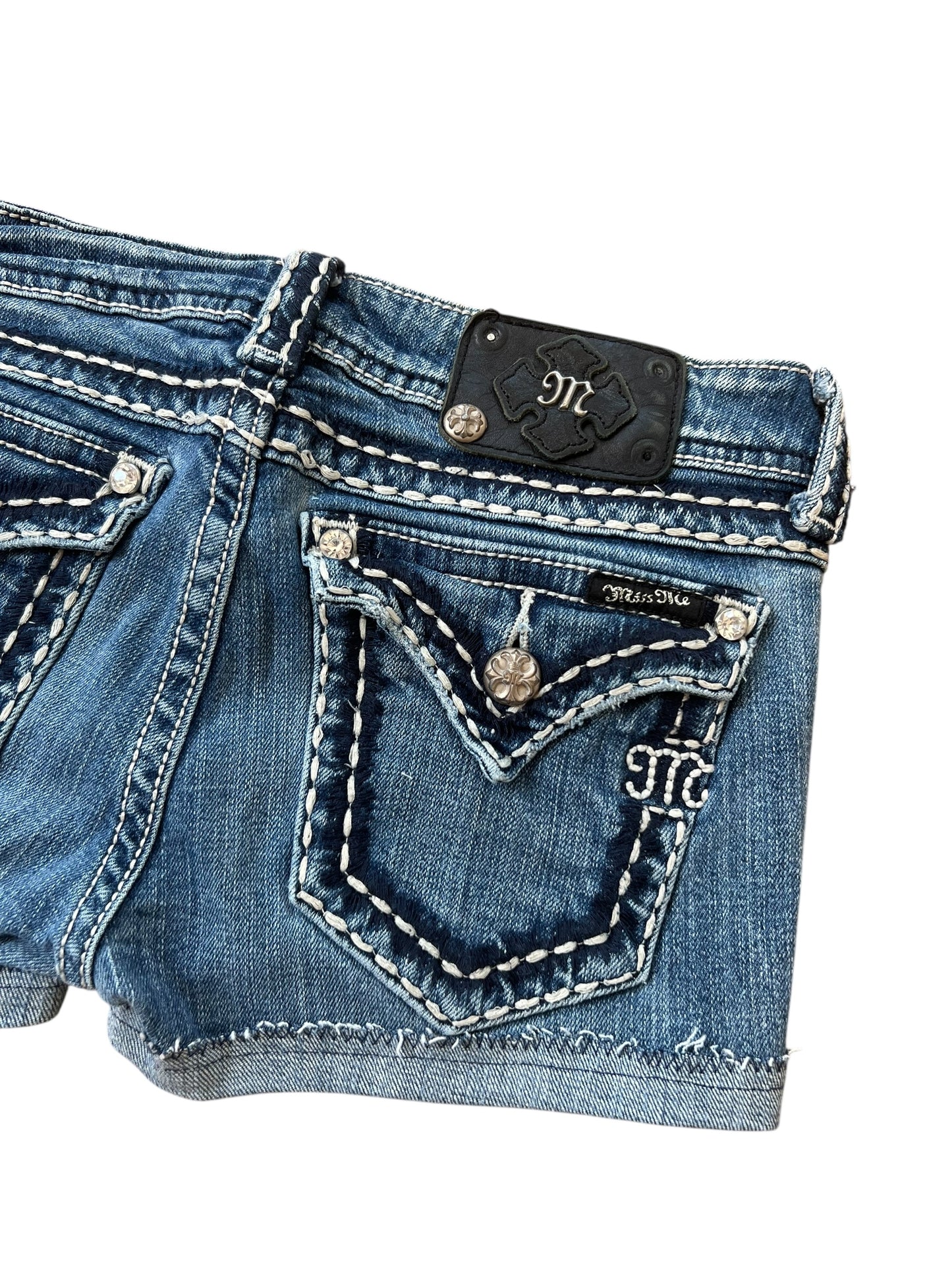 Nahaufnahme der Miss Me Shorts | 27 (S) von ZAZA resell aus blauem Denim mit Bundweite 35, weißen Kontrastnähten, silbernen Knöpfen, einer bestickten Gesäßtasche und einem schwarzen Lederpatch mit M-Logo in der Nähe des Bundes.