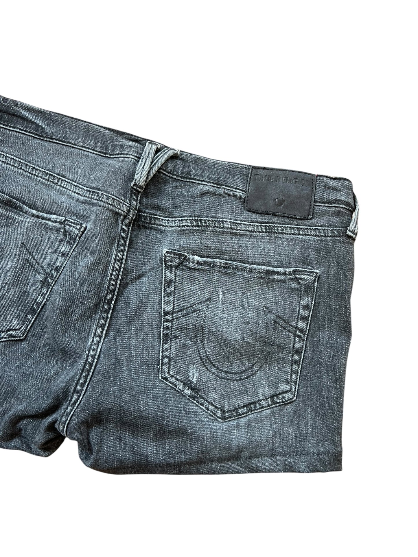 Eine Nahaufnahme der True Religion Shorts | 32 (L) von ZAZA resell zeigt verblassten schwarzen Denim, sichtbare Nähte, eine Gesäßtasche mit einer auffälligen U-förmigen Naht und einen schwarzen Lederaufnäher am Bund.
