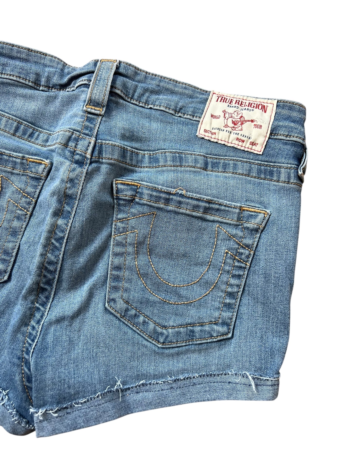 Eine Nahaufnahme der blauen True Religion High Waist Shorts (Größe 30, M) von ZAZA resell mit ausgefranstem Saum, aufgenähtem Hufeisen auf der Gesäßtasche und weißem Markenlabel am Bund.