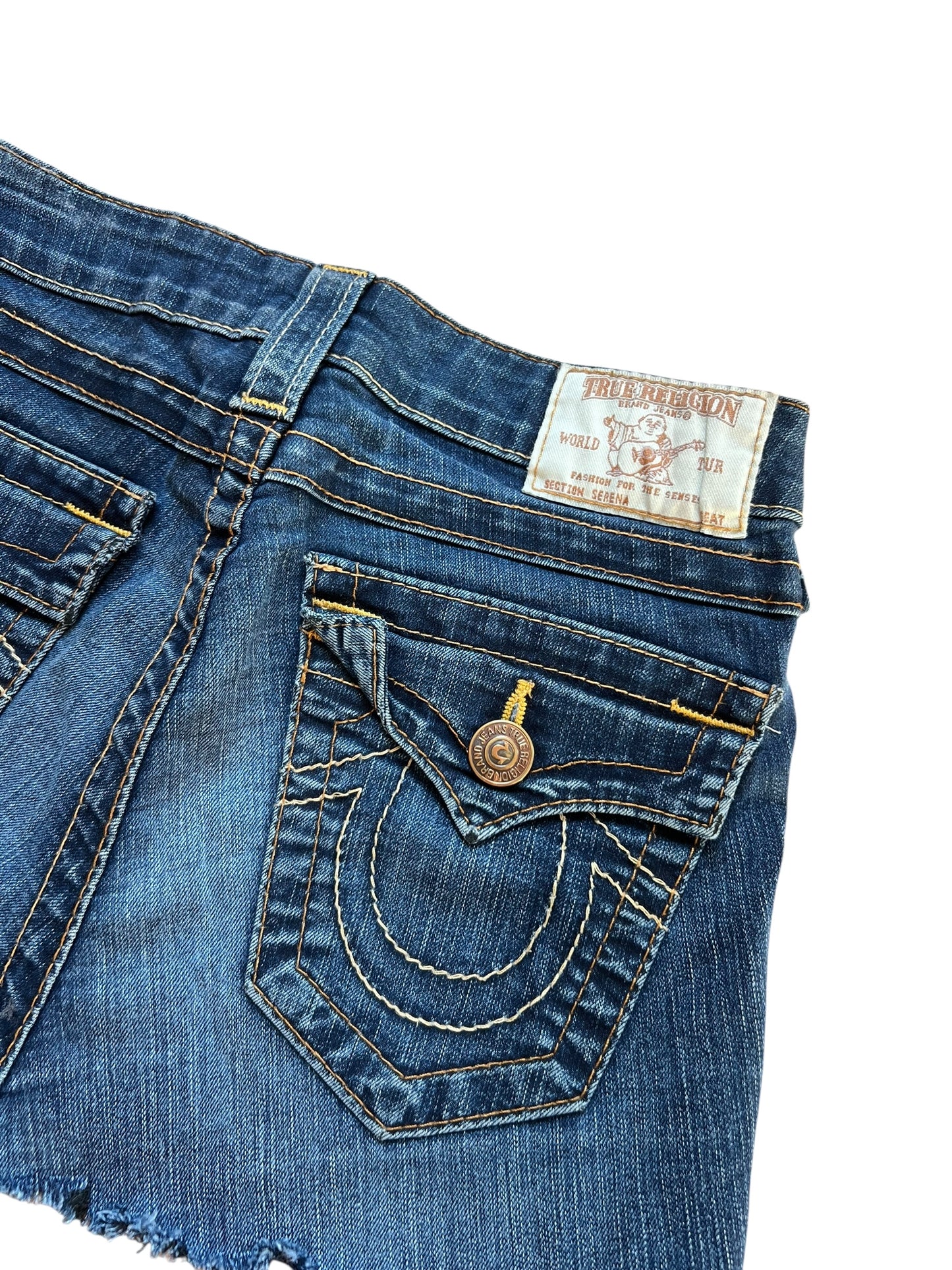 Eine Nahaufnahme eines blauen True Religion Jeansrocks (Größe 24/XS) von ZAZA resell mit gelben Nähten, einem Metallknopf, Gesäßtaschen, einem weißen Markenaufnäher aus Leder über der rechten Tasche und einem ausgefransten Saum. Maße in Zentimetern auf Anfrage erhältlich.