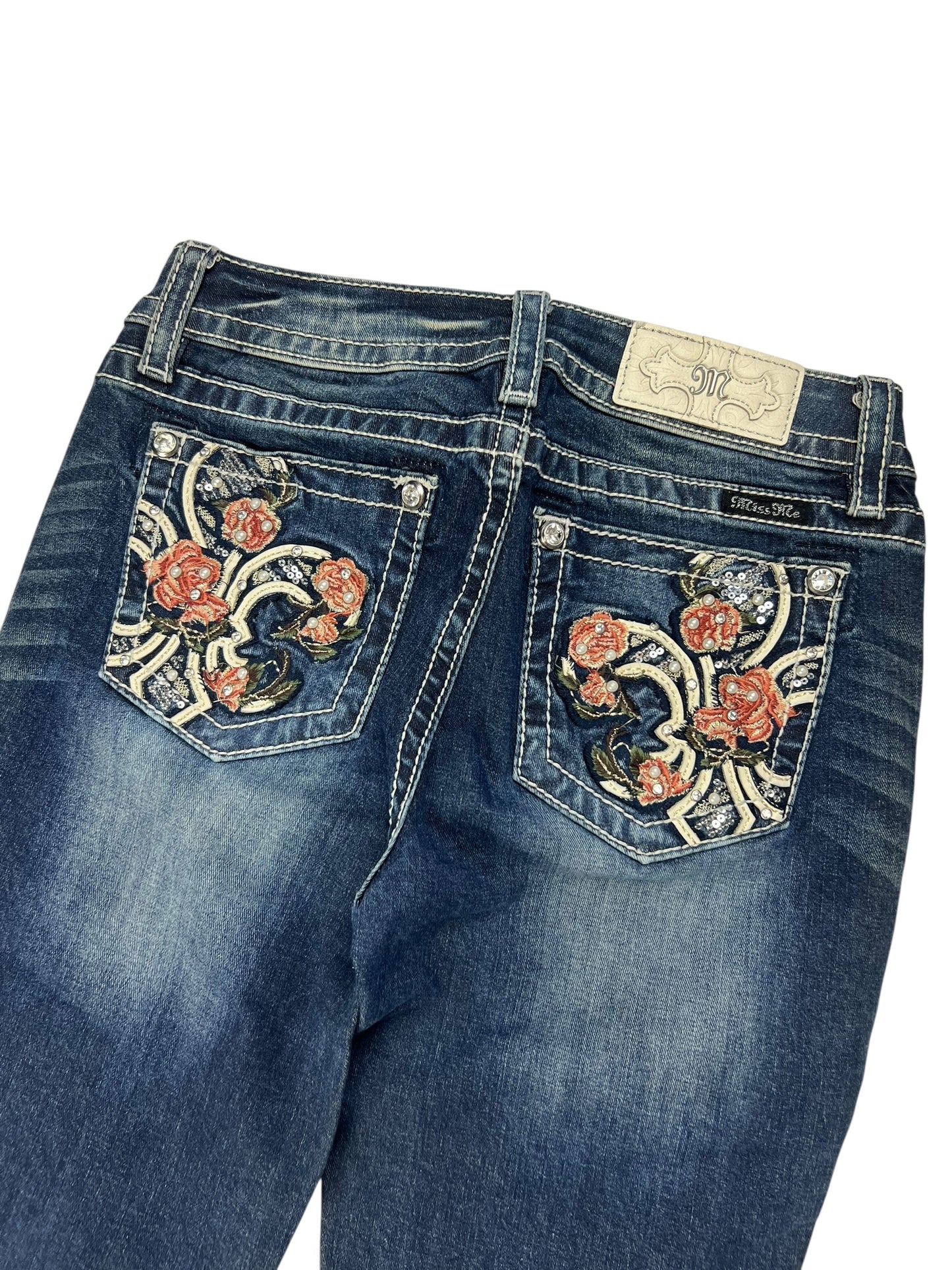 Die Miss Me Low Rise Boot Jeans von ZAZA resell in Größe 27 (S) hat blauen Denim mit ausgeblichenen Details, eine rote und rosa Blumenstickerei auf den Gesäßtaschen, ein weißes Markenpatch aus Leder am Bund und eine Passform, die als Außenbeinlänge und Bundweite für Größe 27 angegeben ist.