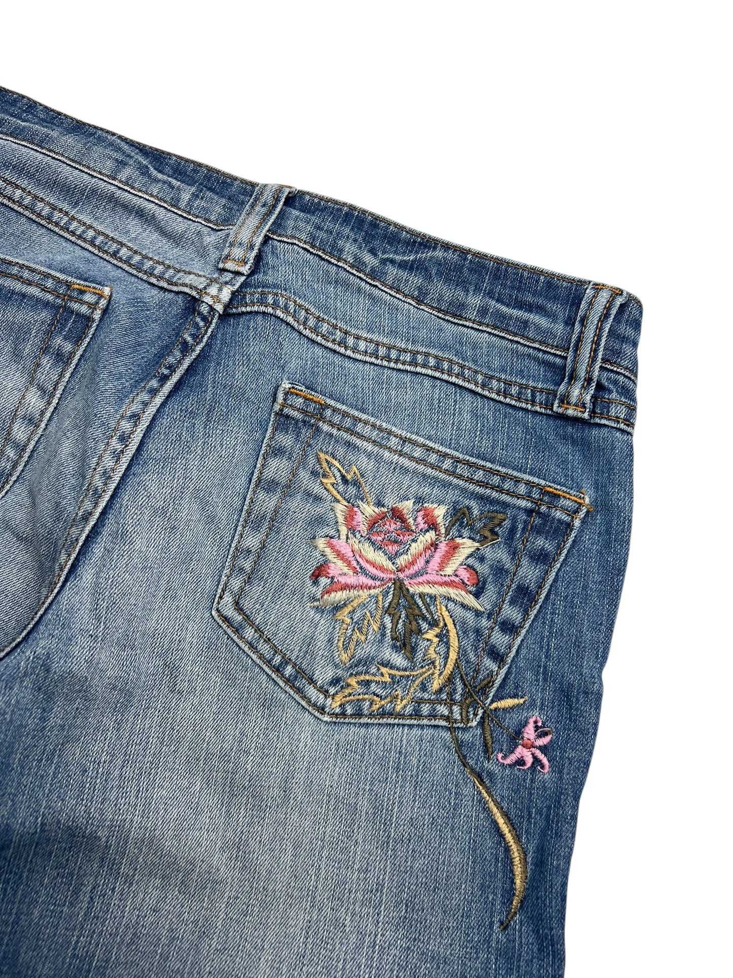 Eine Nahaufnahme der Y2K Low Rise Boot Jeans (W26 x L30) von ZAZA Resell zeigt ein gesticktes rosa und grünes Blumenmuster auf der Gesäßtasche, sichtbare Nähte, verblasste Details und einen Blick auf die Bundweite als Passformreferenz.