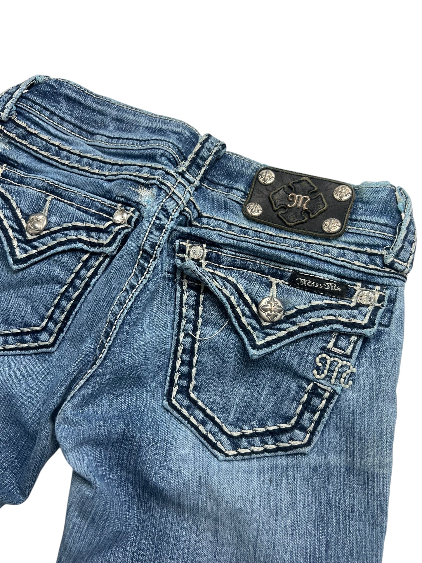 Eine Nahaufnahme der Rückseite der Miss Me Low Rise Boot Jeans (W25 x L30) von ZAZA Resell, mit weißen Nähten, silbernen Knöpfen, verzierten Taschen und einem schwarzen Markenaufnäher mit Logo, alles in einem trendigen Used-Look.