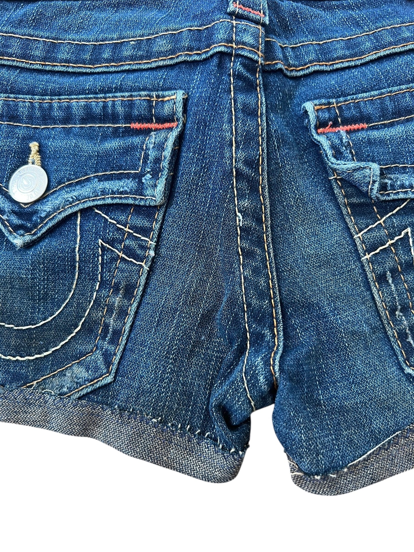 Nahaufnahme der Rückseite der True Religion Shorts | 23 (XS) von ZAZA resell aus blauem Denim, mit zwei Taschen, weißen Nähten, Metallknöpfen, roten Fadenakzenten, gerolltem Saum und einer abgenutzten Textur für einen authentischen Cowboy-Look.