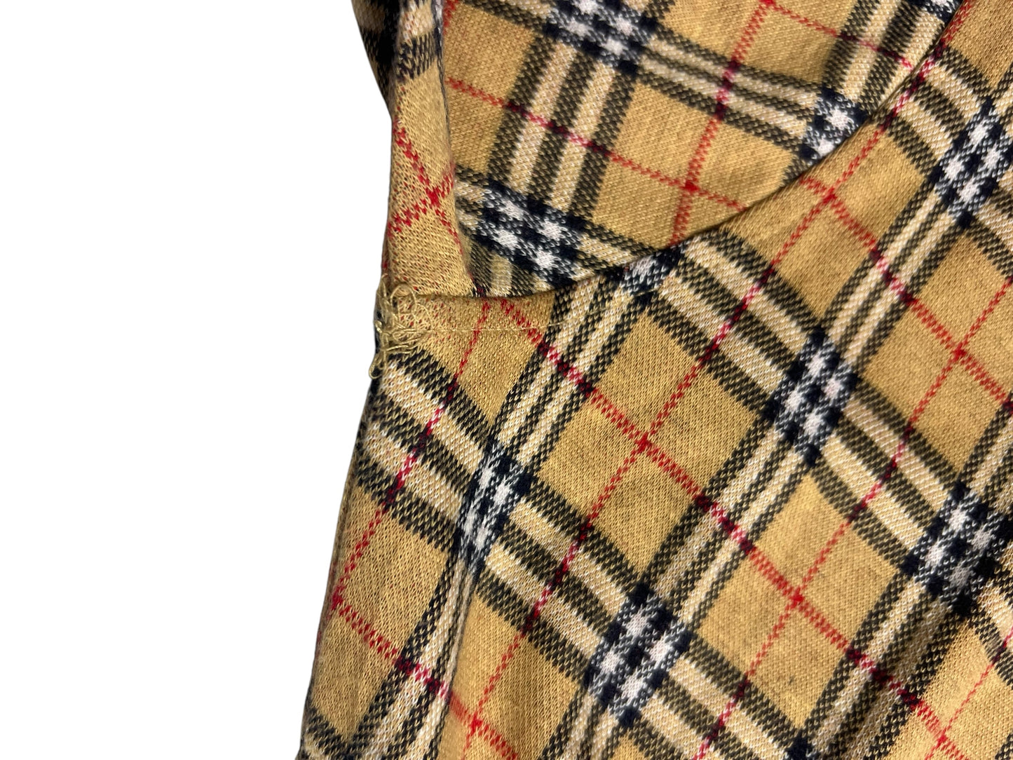 Nahaufnahme eines hellbraunen Burberry-Poloshirts (Größe S) mit einem karierten Muster aus schwarzen, weißen und roten Linien. Das Bild zeigt eine Naht und eine Falte, die auf Kleidungsdetails hinweisen. Marke: ZAZA resell. Zustand 9/10.