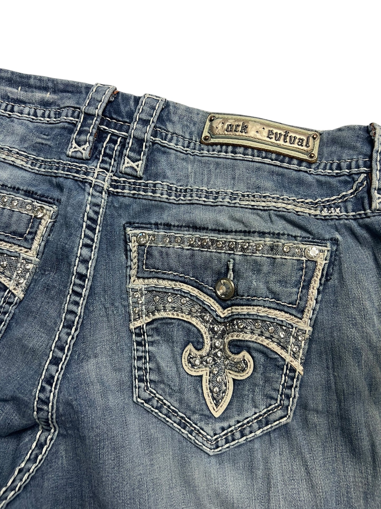 Eine Nahaufnahme der blauen Jeans von ZAZA resell, Rock Revival Mid Rise Boot | 32 (XL), zeigt Ziernähte, Strasssteine, eine Pattentasche mit Knopf und ein Rock Revival Patch aus Leder. Bundweite 46 und Außenbeinlänge 110 cm sorgen für perfekten Sitz und einzigartigen Style.