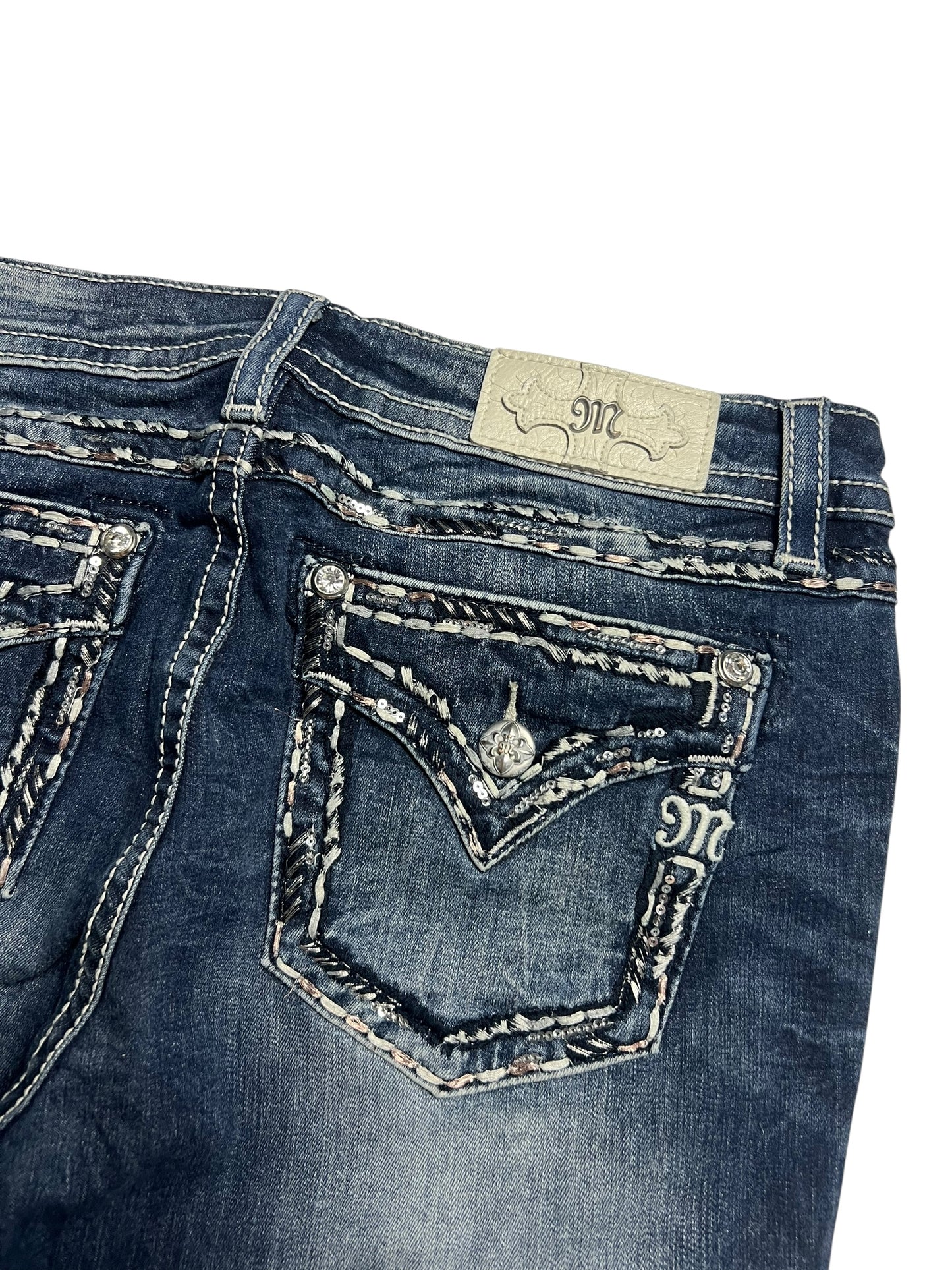 Eine Nahaufnahme der Miss Me Low Rise Boot | 32 (L) Jeans von ZAZA resell. Sie zeigt dunkelblauen Denim, weiße und graue Nähte, Gesäßtaschen mit Klappe und Knöpfen, einen hellen Markenaufnäher aus Leder in der Nähe des Bunds und eine durchsichtige Bundweite zur Größenbestimmung.