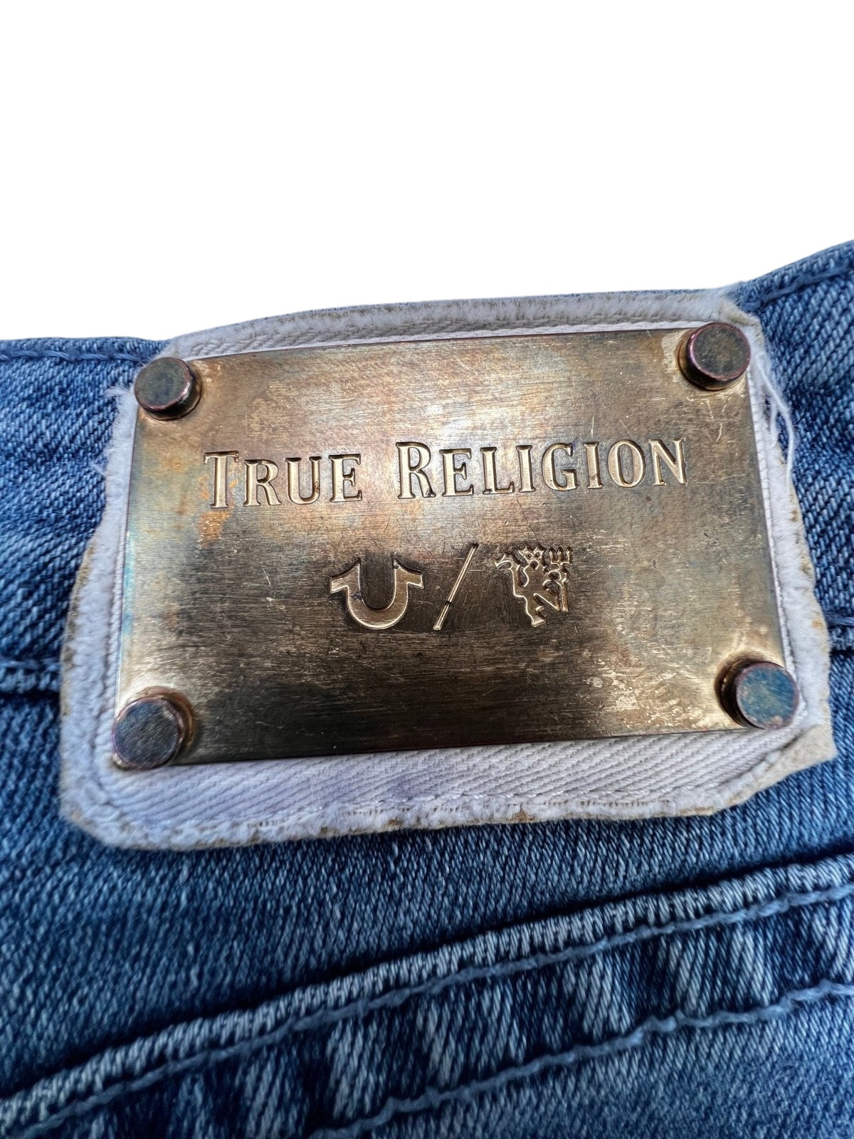 Nahaufnahme eines metallischen Etiketts auf einer Blue-Denim-Jeans mit der Gravur „ZAZA resell“ und vier Nieten. Diese seltene True Religion x Manchester United Mid Rise Flare (W34 x L30) trägt die Markenlogos. Zustand: 9/10. Maße auf Anfrage.