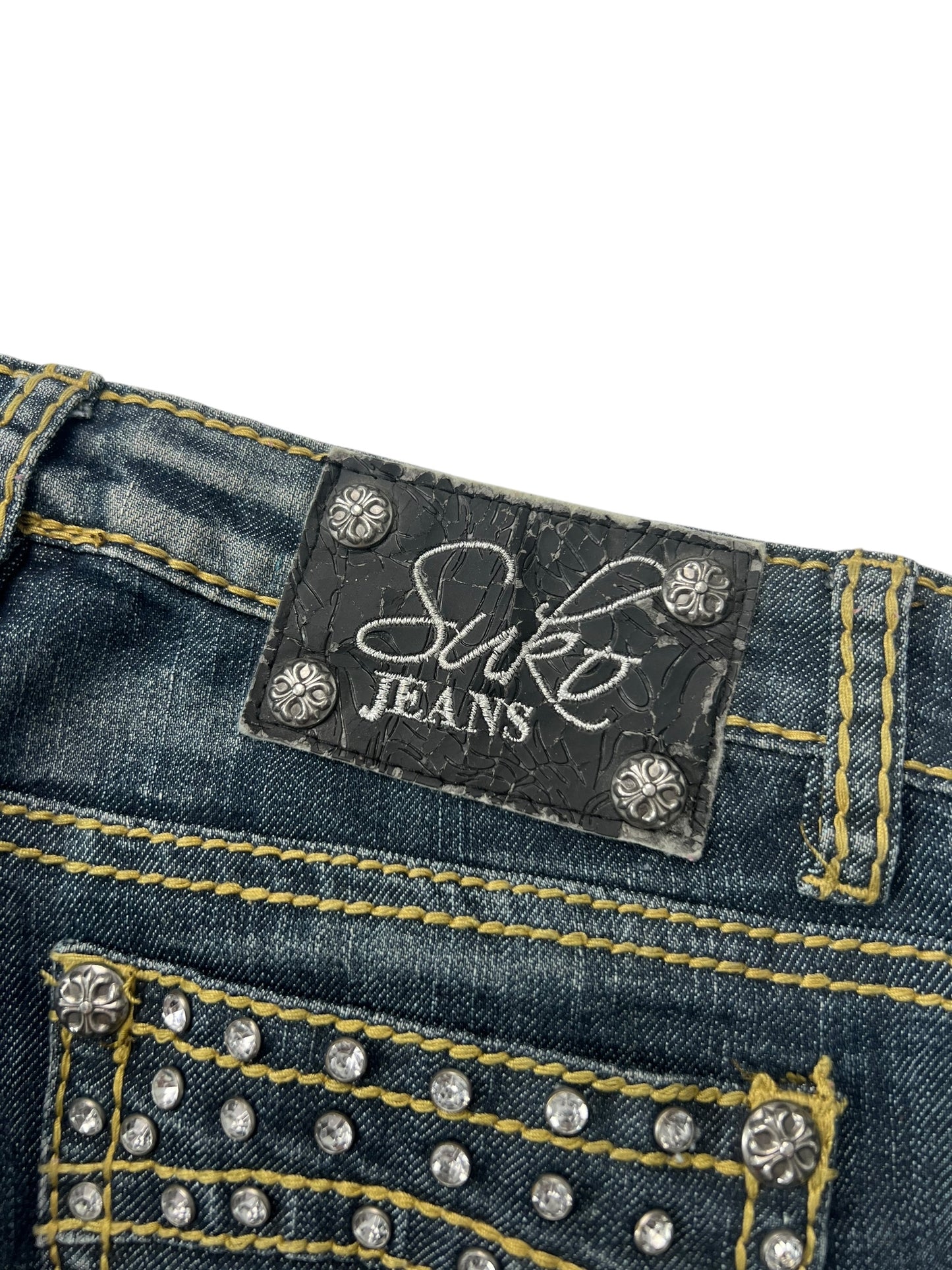 Eine Nahaufnahme des hinteren Hosenbunds des Suko Jeans Mid Rise Boot (W26 x L34) von ZAZA Resell zeigt blauen Denim mit gelben Nähten, silbernen Nieten und einem schwarzen Lederaufnäher mit der Aufschrift „Suko JEANS“ in weißer Schrift und Blockbuchstaben.