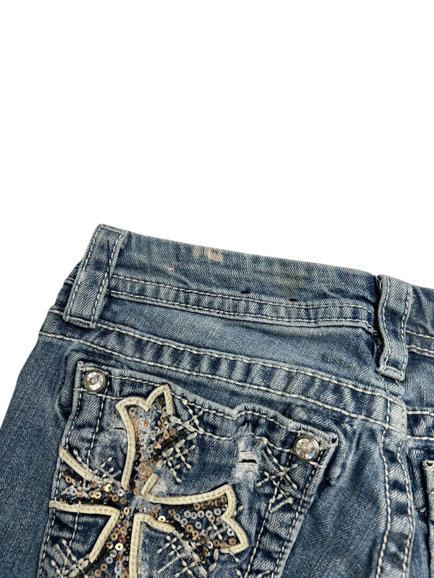 Nahaufnahme der Miss Me Mid Rise Boot Jeans (W28 x L26) von ZAZA Resell. Sie besteht aus blauem Denim mit einem gestickten und mit Pailletten besetzten Kreuz auf der Gesäßtasche, sichtbaren Nähten, silbernen Knöpfen und einem Labelgröße-Etikett.