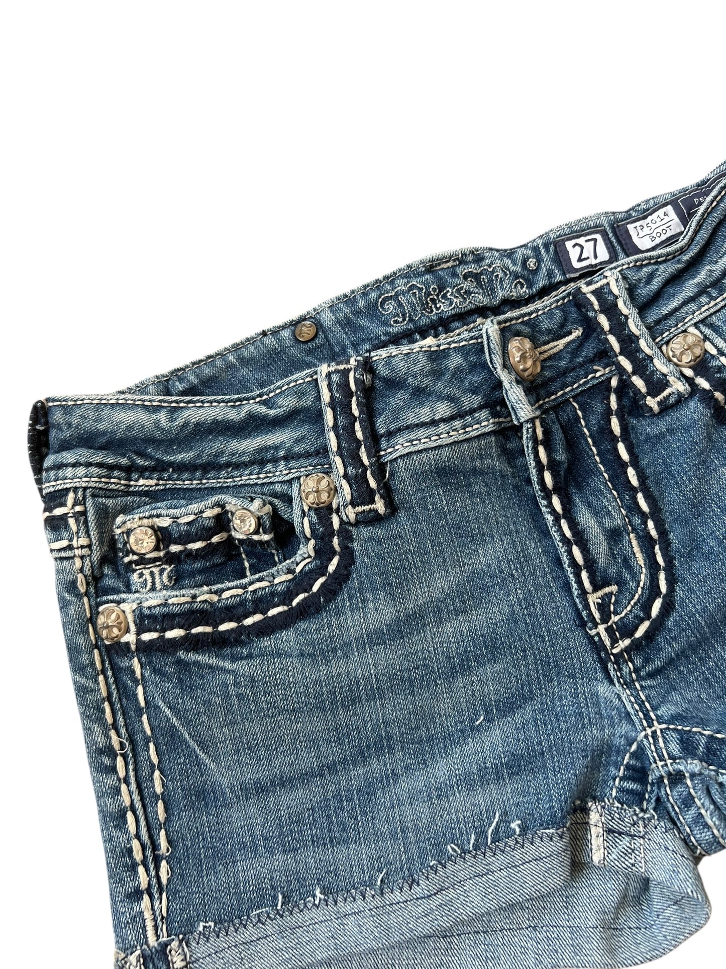 Nahaufnahme der blauen Denim Miss Me Shorts | 27 (S) von ZAZA resell, mit weißen Nähten, Ziernieten, einem sichtbaren Markenetikett am Bund und dem Größenetikett Labelgröße 27 mit Bundweite 35.