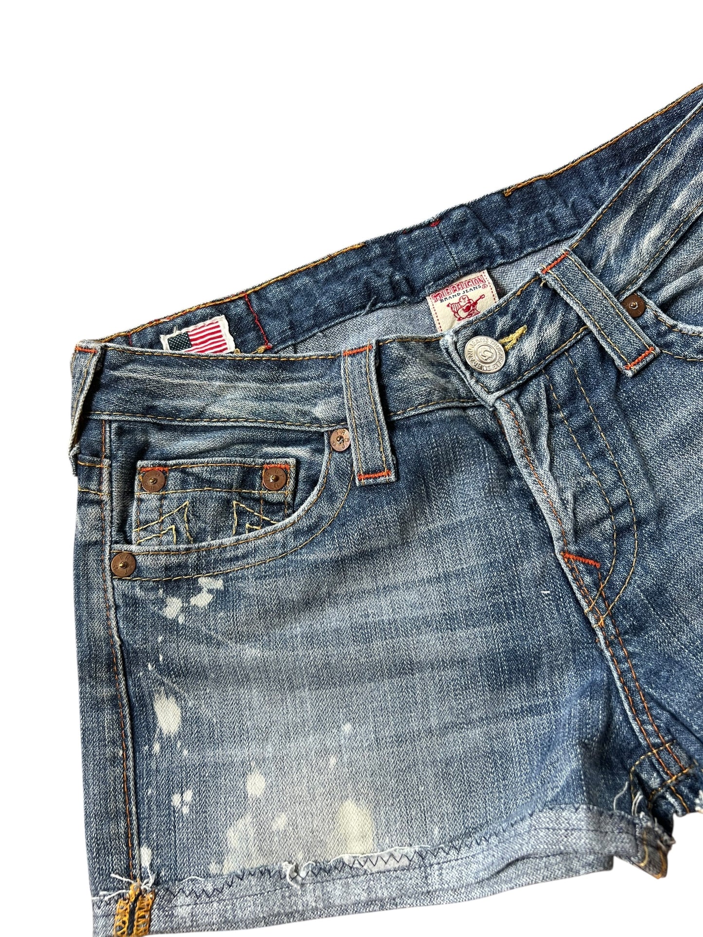 Eine Nahaufnahme der True Religion Shorts | 27 (M) mit Bundweite 39 von ZAZA resell, mit Distressed Denim, verblassten Bereichen, Farbspritzern, freiliegenden Nähten, einer Vordertasche, Gürtelschlaufen und einem sichtbaren Knopfverschluss.