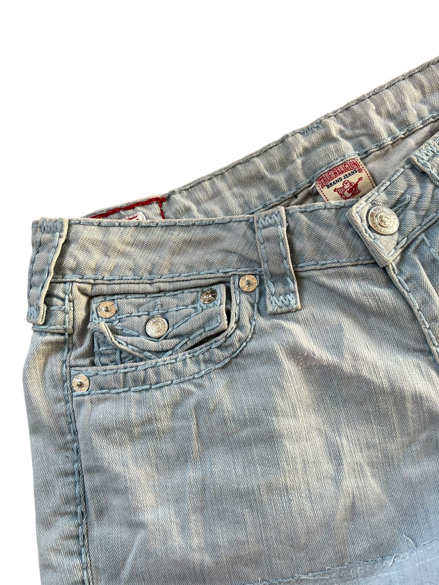 Nahaufnahme der True Religion Shorts von ZAZA resell aus hellblauem Denim, mit sichtbaren Nähten, Metallnieten an der Vordertasche, einer kleinen Münztasche, Gürtelschlaufen und einem teilweise sichtbaren Markenetikett im Bund. Maße in Zentimetern. Größe: 30 (M).