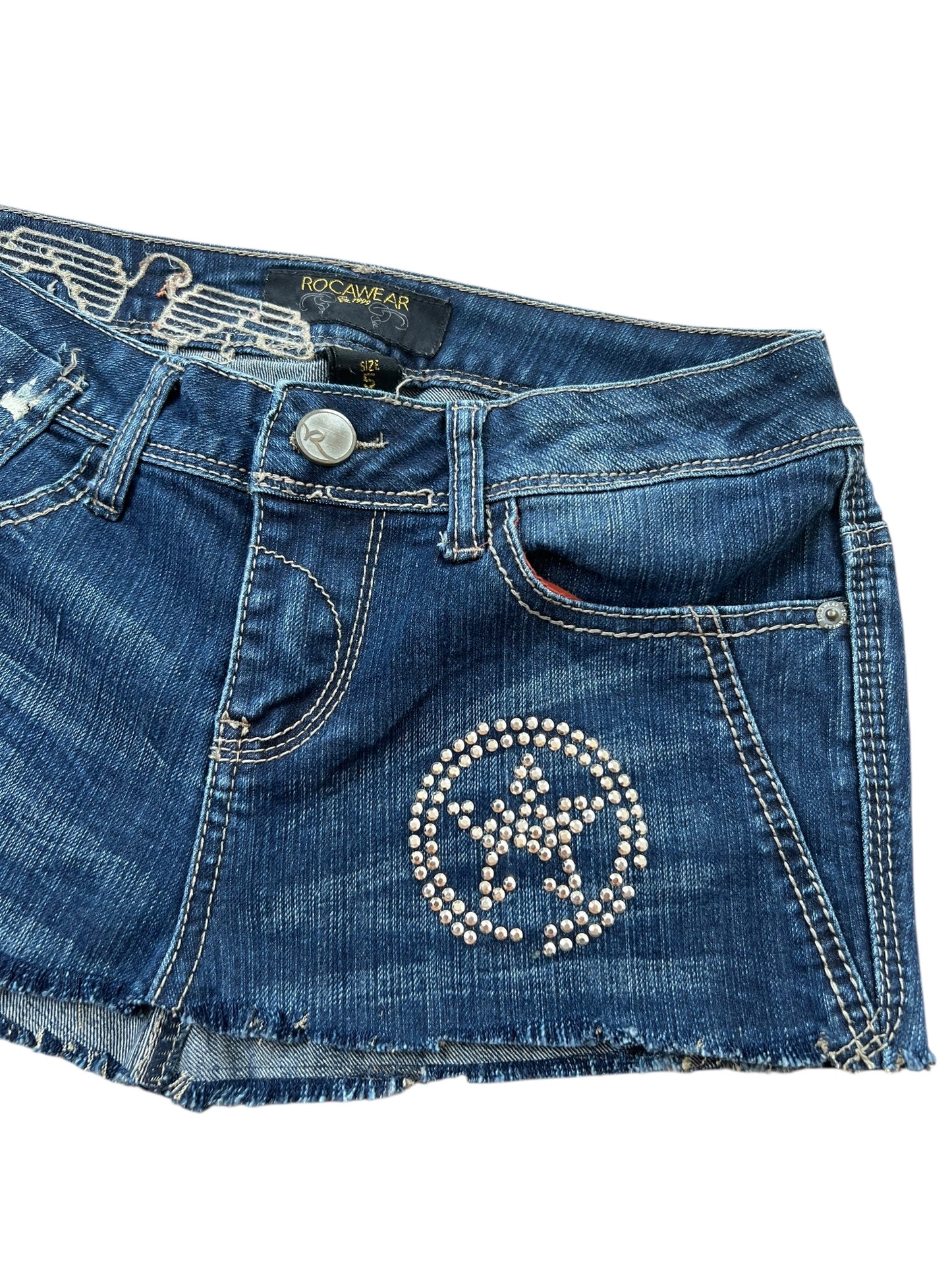 Rocawear Rock | W27 von ZAZA resell: Jeans-Minirock mit ausgefranstem Saum, Fronttaschen, Gürtelschlaufen und silbernem Stern-Kreis-Muster auf der rechten Seite. Bundweite sorgt für eine tolle Passform. Label: Rocawear.