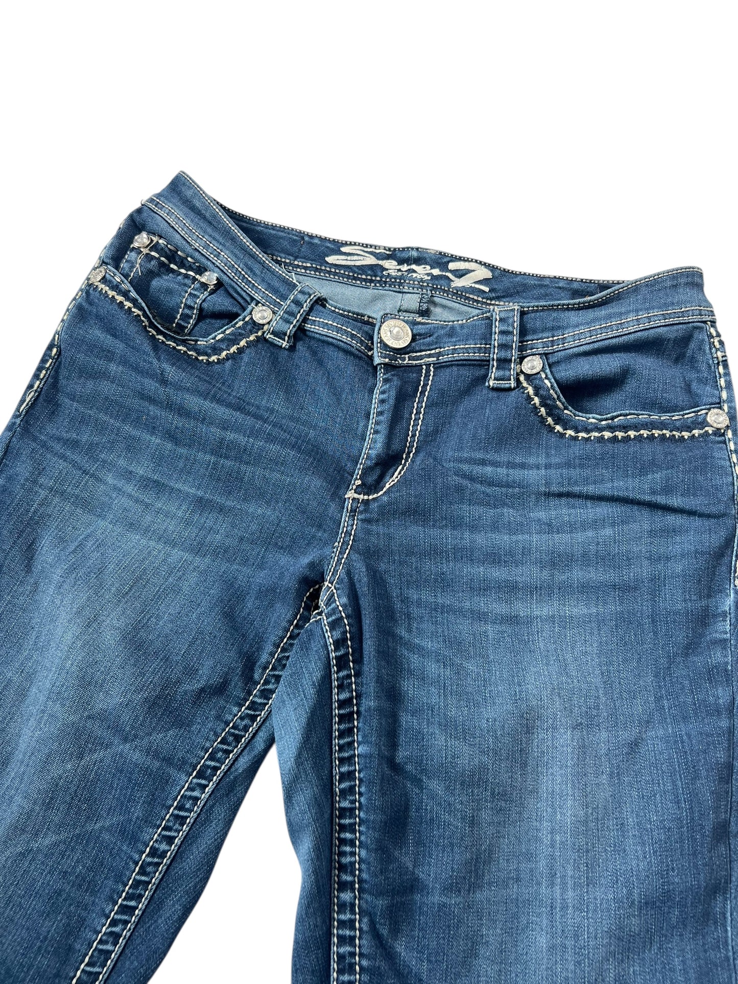 Die von ZAZA weiterverkauften Seven Mid Rise Boot Jeans in W33 x L34 bestehen aus blauem Denim mit weißen Kontrastnähten, silbernen Nieten und einem Knopfverschluss, der Bundweite und Beinöffnung hervorhebt und flach auf weißem Hintergrund liegt.