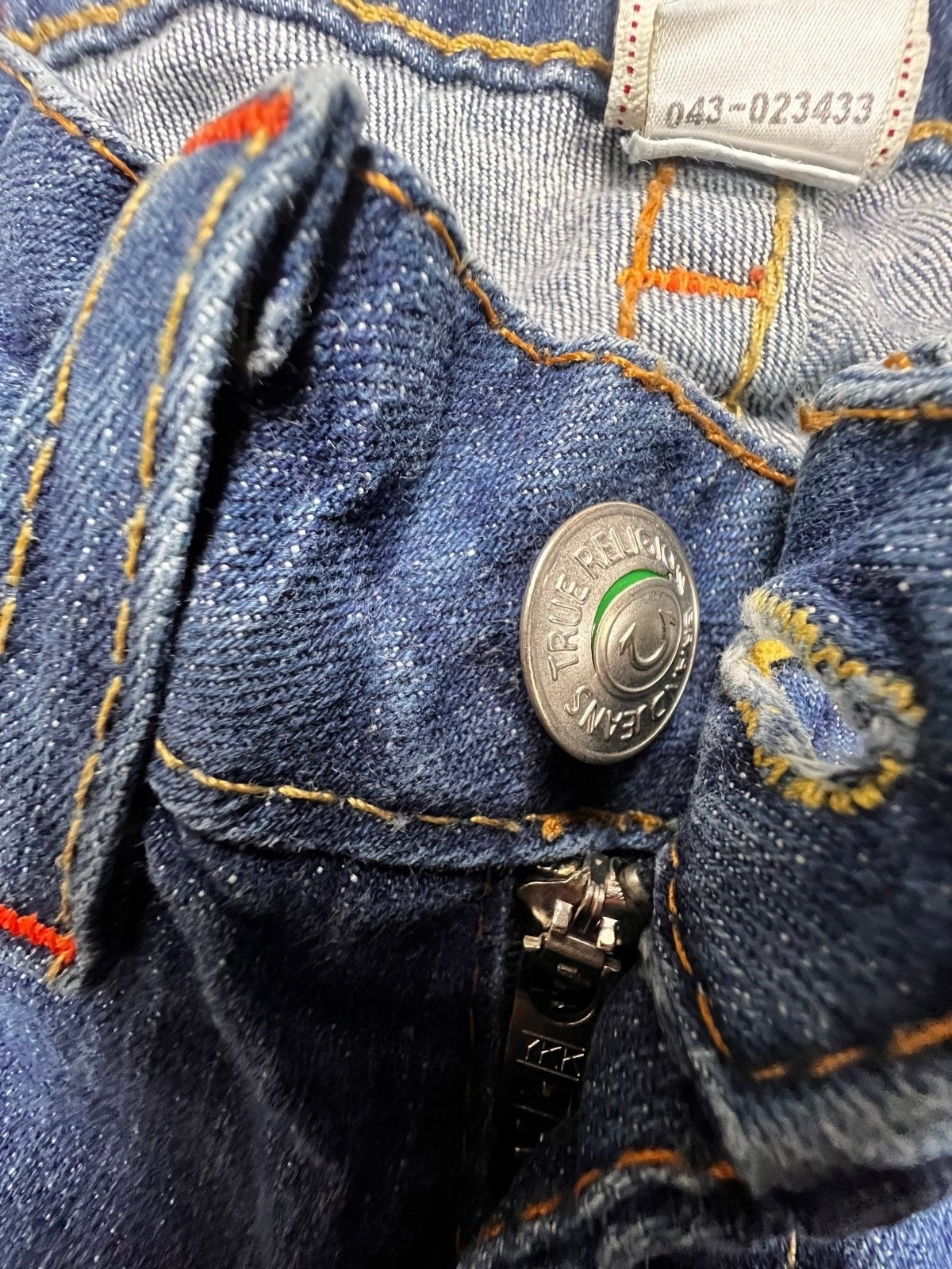 Nahaufnahme der True Religion Johnny – Low Rise Straight Jeans (W24 x L30) von ZAZA Resell: blauer Denim, Metallknopf mit Logo, sichtbarer Reißverschluss, orangefarbene Nähte und weißes Innenetikett mit Größenangaben in Zentimetern und Zahlen.