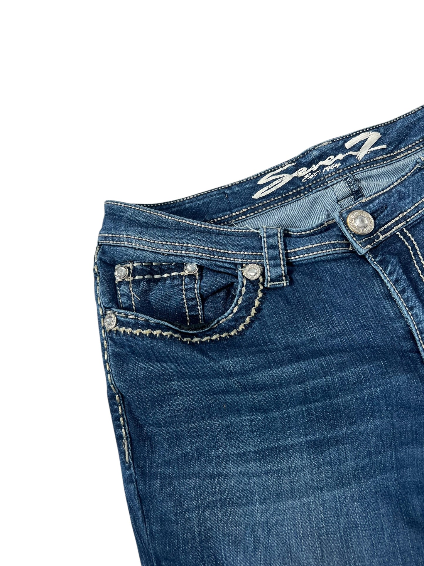 Eine Nahaufnahme der von ZAZA weiterverkauften Seven Mid Rise Boot Jeans (W33 x L34). Sie präsentiert sich in blauem Denim, mit weißen Nähten, dekorativen Taschendetails, einem Metallknopf, einem Teil des Markenetiketts am Bund und einer klaren Labelgröße zur genauen Größenbestimmung.