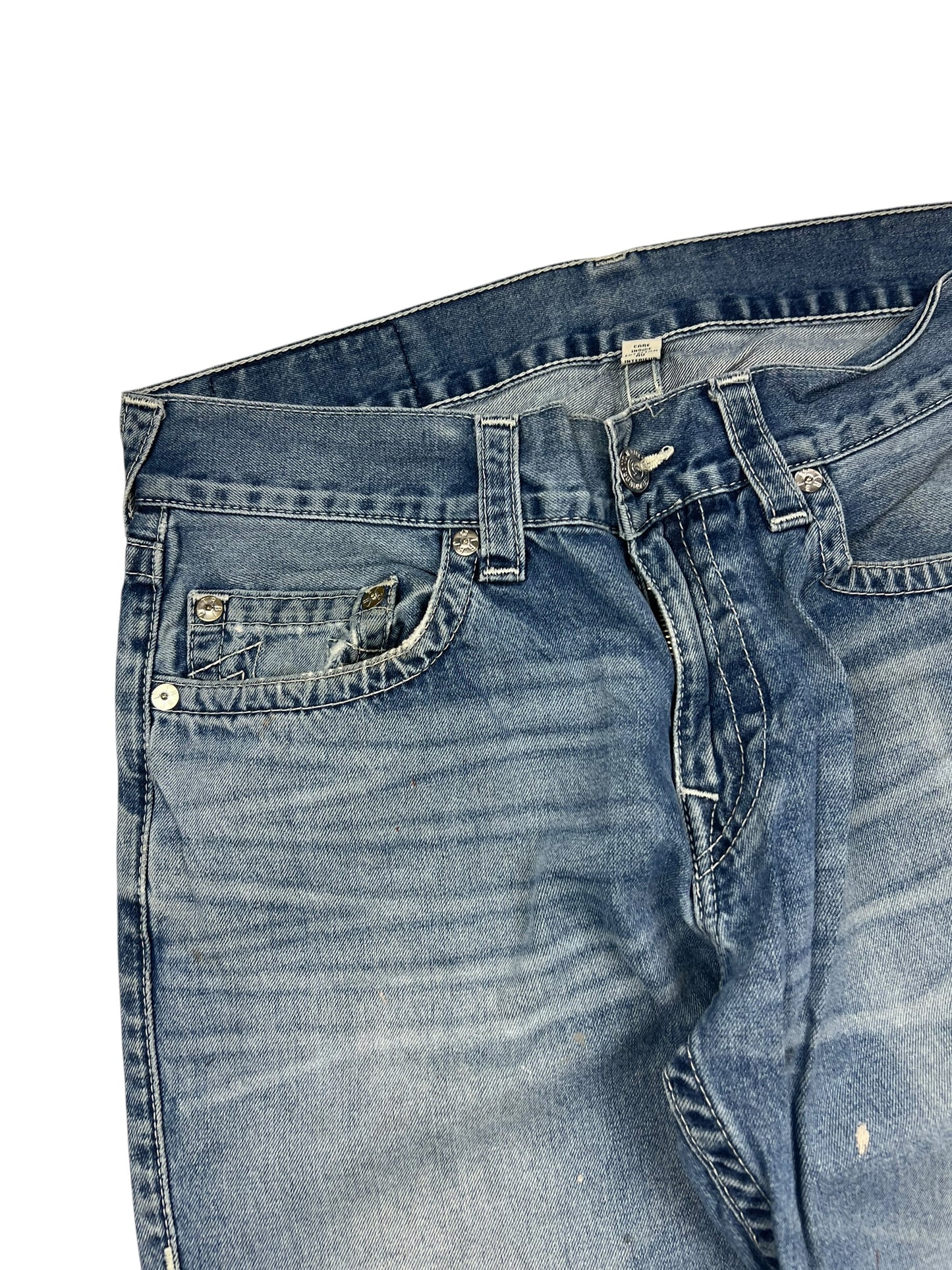Eine Nahaufnahme der True Religion Straight | 34 (L) Blue Denim Jeans von ZAZA resell zeigt abgenutztes, verblasstes Gewebe, sichtbare Taschen, Gürtelschlaufen, einen Metallknopf und die Etikettengröße in der Nähe des Bundes auf weißem Hintergrund.
