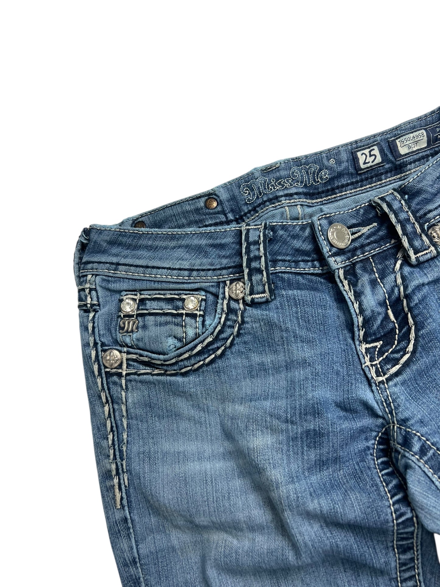 Eine Nahaufnahme der Miss Me Low Rise Boot Jeans von ZAZA Resell in W25 x L30, mit blauem Denim mit weißen Kontrastnähten, dekorativen silbernen Vordertaschenknöpfen, genähten Details und einem Bundetikett mit der Größenangabe.