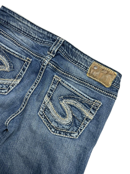 Eine Nahaufnahme der Silver Jeans Low Rise Boot von ZAZA resell in Größe 29 (M). Sie zeigt blauen Denim mit aufgestickten Taschendesigns, einen braunen Leder-Markenaufnäher am Bund und Bundweite-Details für eine perfekte Passform.