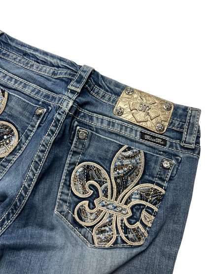 Eine Nahaufnahme der blauen Denim-Jeans Miss Me Mid Rise Boot (Größe 30/M) von ZAZA resell, mit einem verzierten Fleur-de-Lis-Strassstein-Design auf der Gesäßtasche und einem dekorativen Metall-Label über der Tasche für eine perfekte Passform.