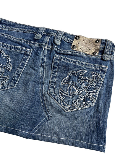 Eine Nahaufnahme des Miss Me Skirt | 28 (M) von ZAZA resell zeigt blauen Denim mit weiß bestickten Gesäßtaschen und einem Metallic-Patch in der Nähe des Bundes, der Details wie Bundweite und Außenbeinlänge für eine individuelle Rock-Passform hervorhebt.